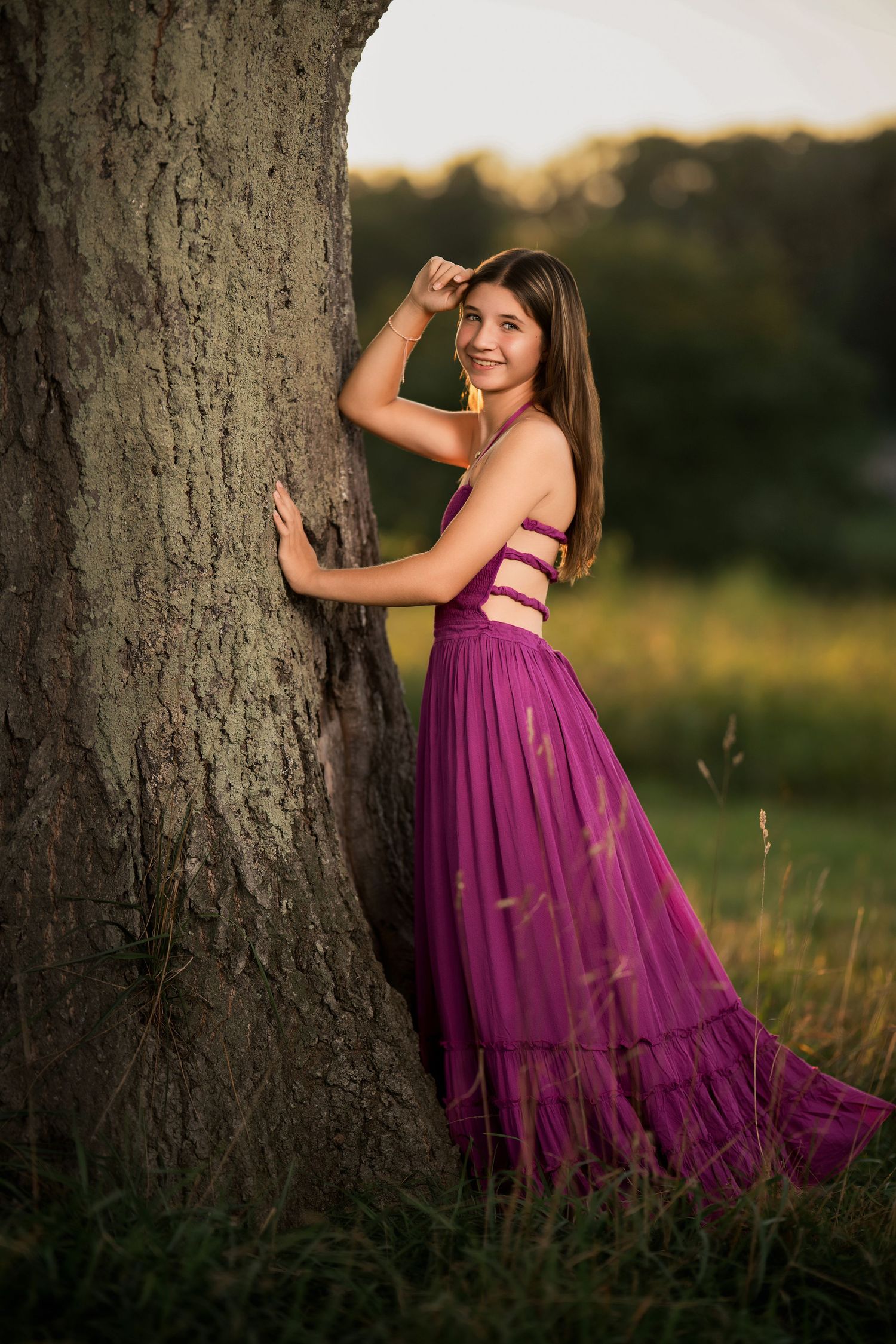 Young woman in purple maxi dress leaning against large tree trunk in golden hour sunlight outdoors.