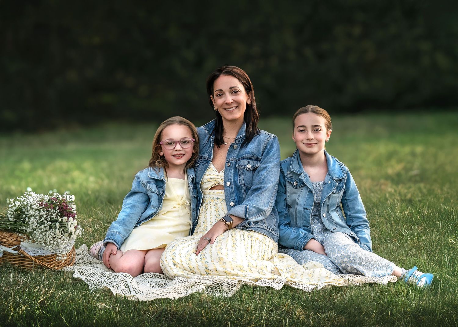 A smiling woman and two young girls in denim jackets sit on a lace blanket in a green grassy field.