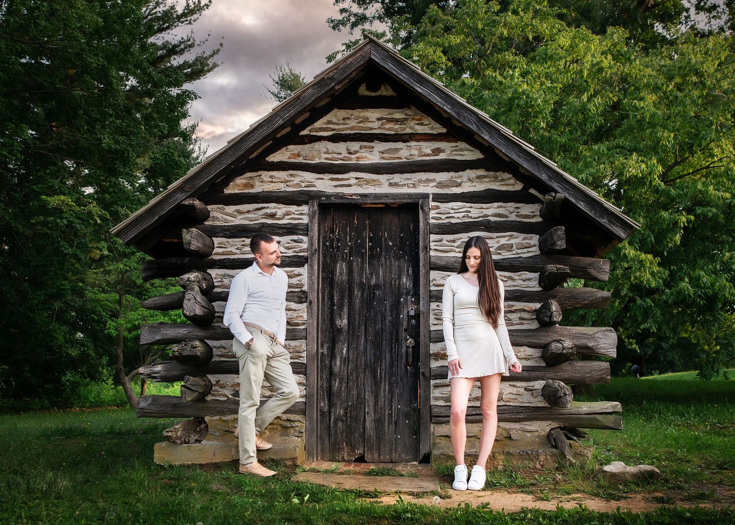Couple posing by rustic log cabin with dark wooden door surrounded by lush green trees.