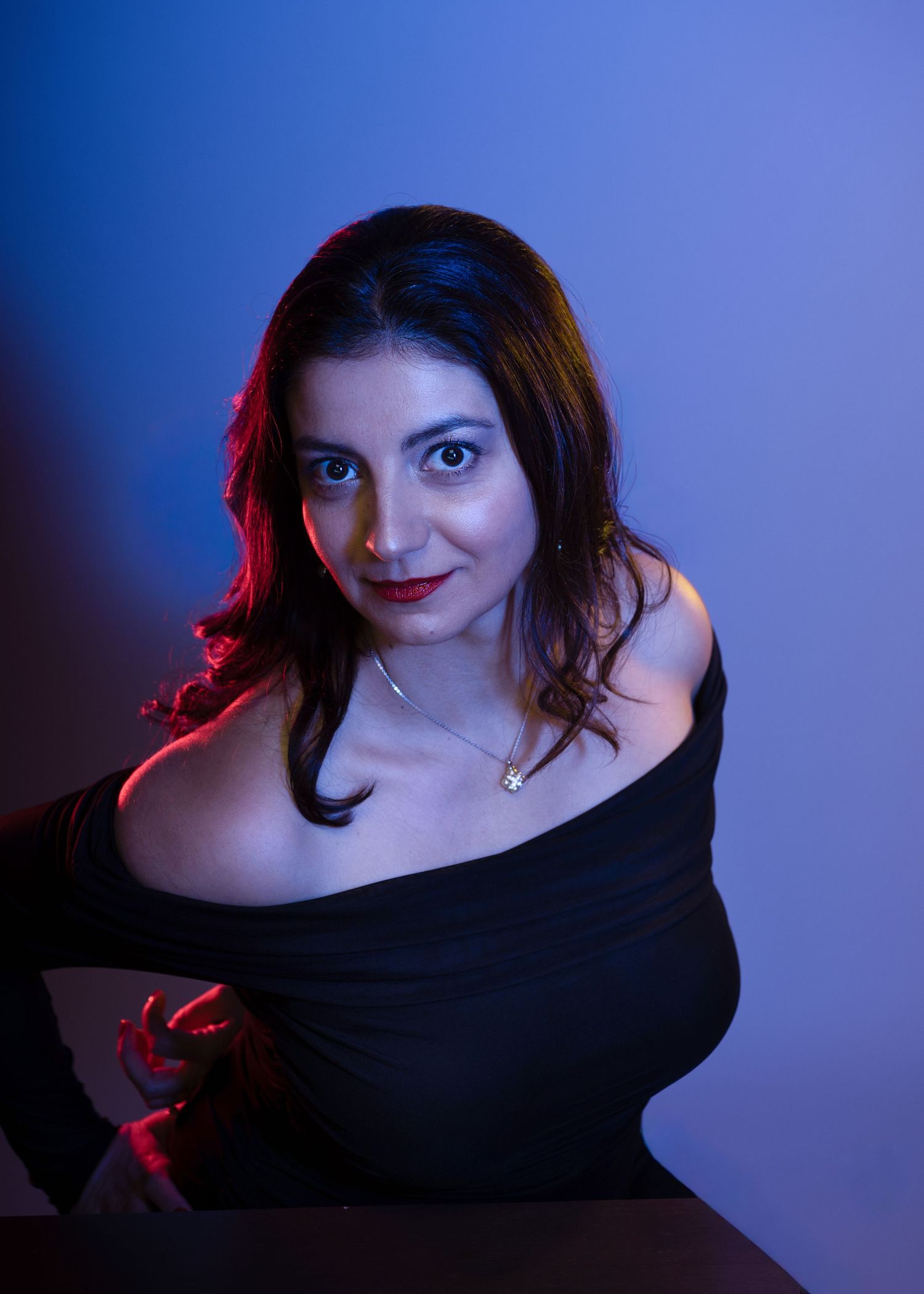 Woman in black off-shoulder top with red lips poses against blue backdrop with dramatic red and blue lighting.