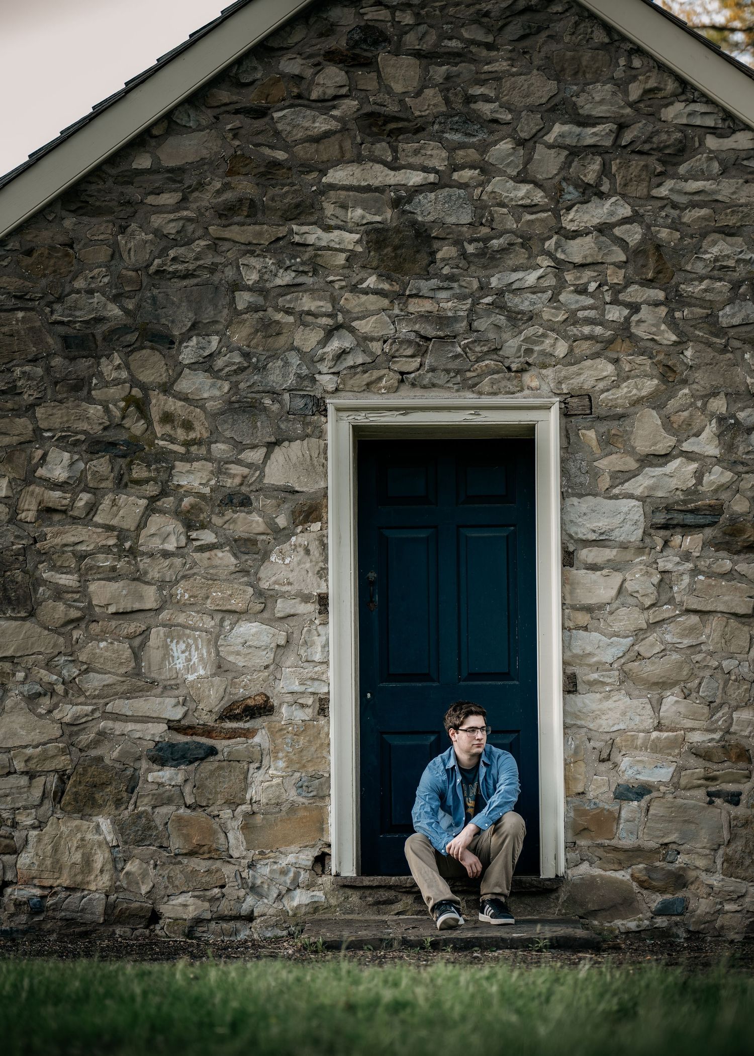 Young man with glasses sitting on steps of a rustic stone building with a blue door.