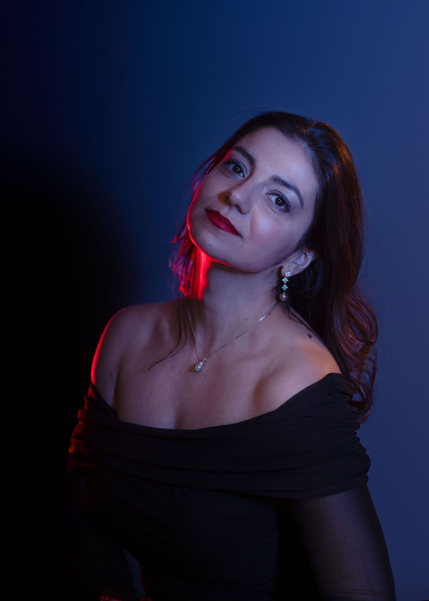 Woman in black off-shoulder top with red lip and pearl earrings posed against dark blue background with dramatic red lighting.