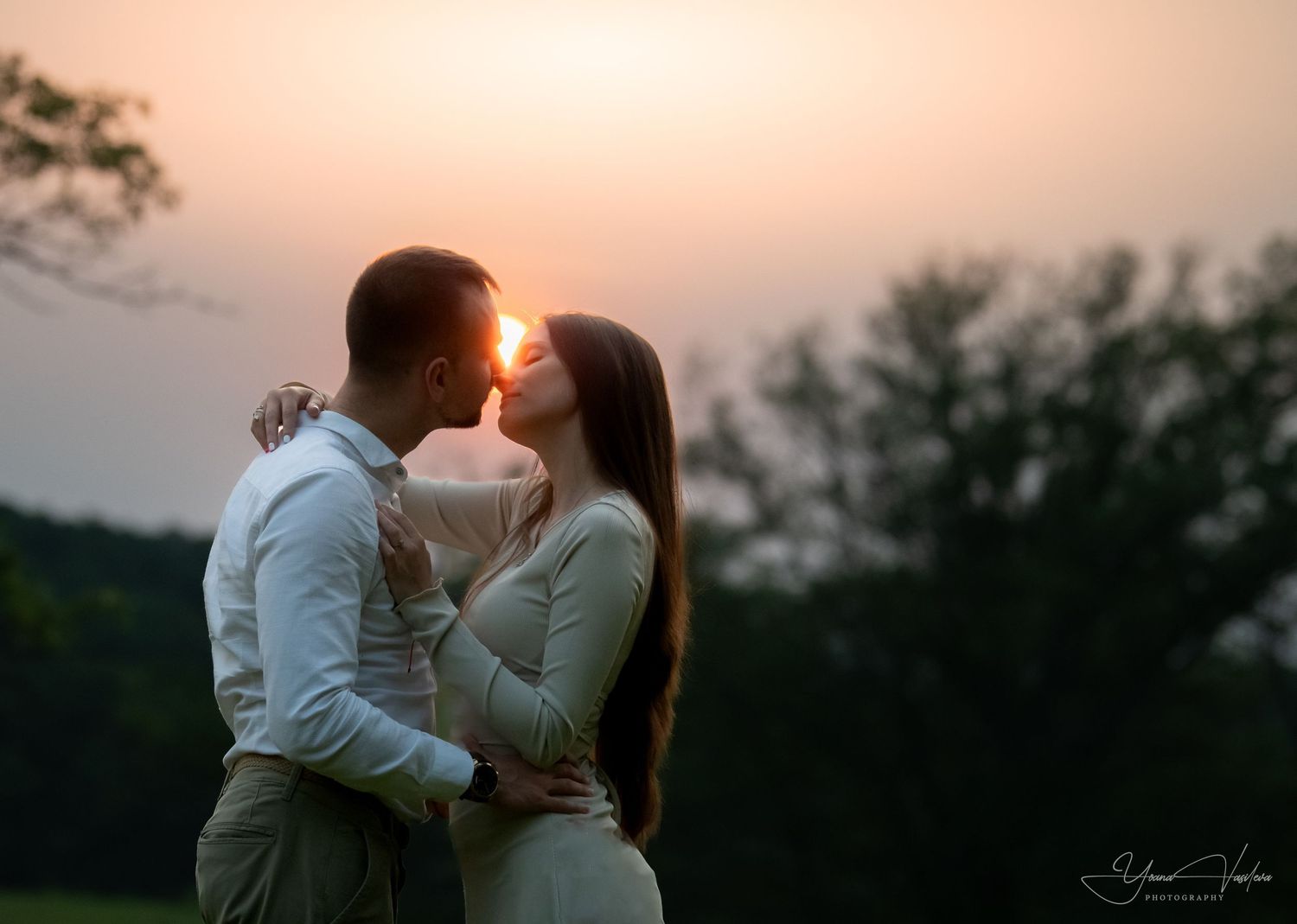 Couple sharing a romantic sunset kiss outdoors with golden light glowing behind them in a lush green landscape.