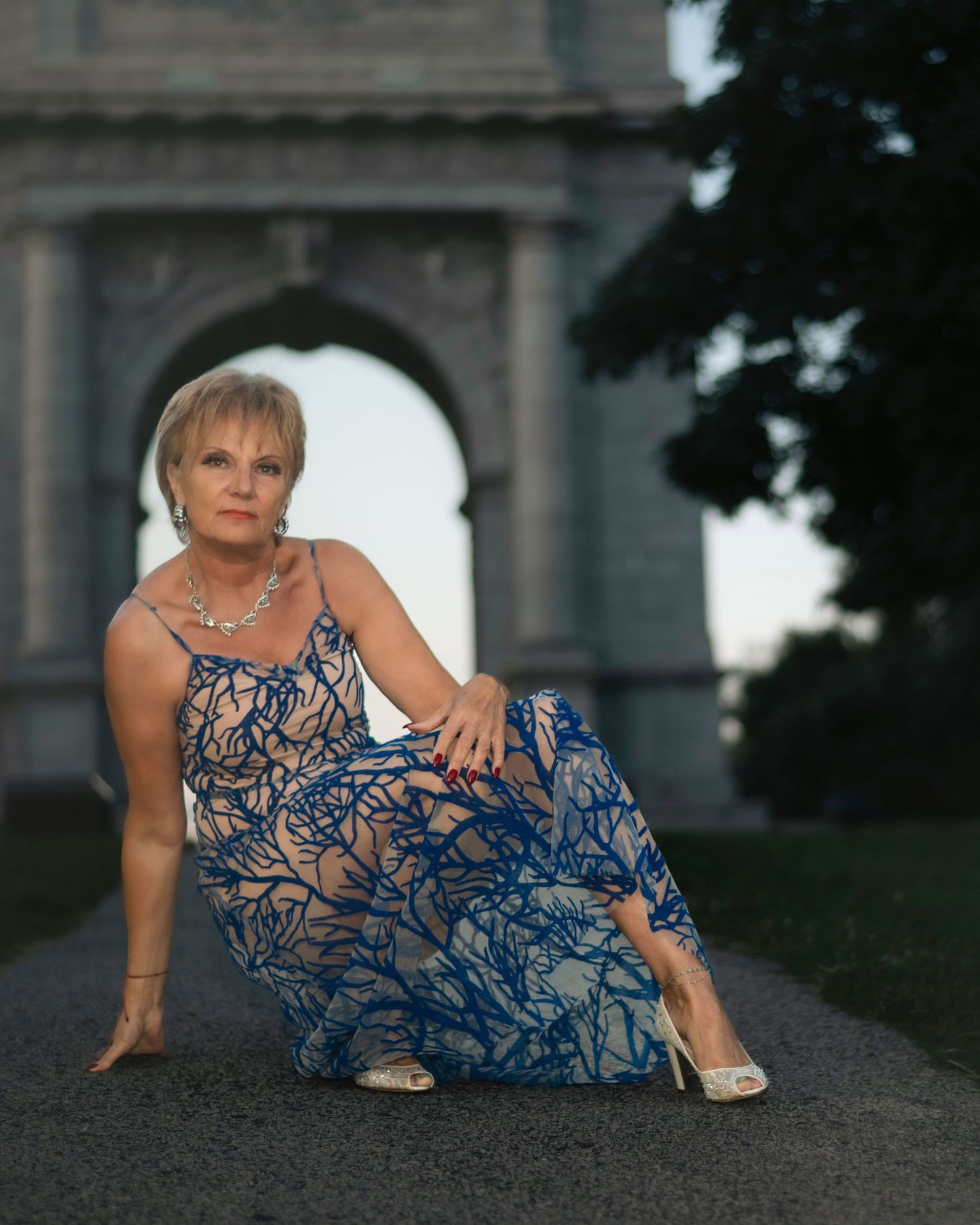 Woman in blue floral maxi dress posing before stone arch monument at dusk during her outdoor lifestyle photography session on the Main Line.