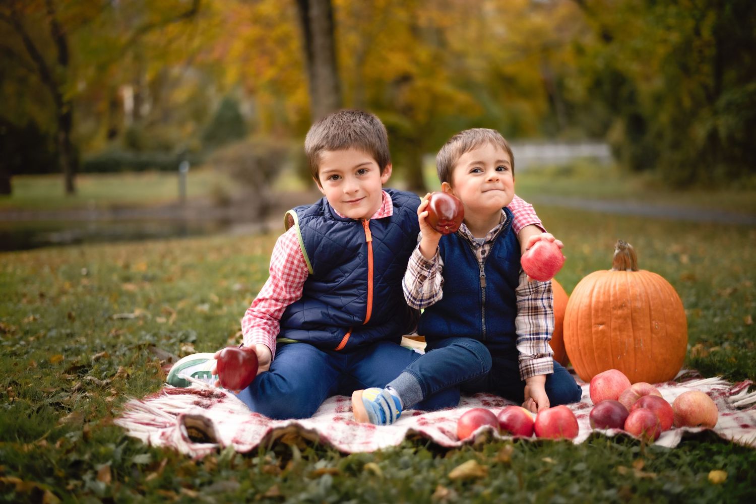 Children sitting on a blanket with pumpkins and autumn leaves in a park setting.