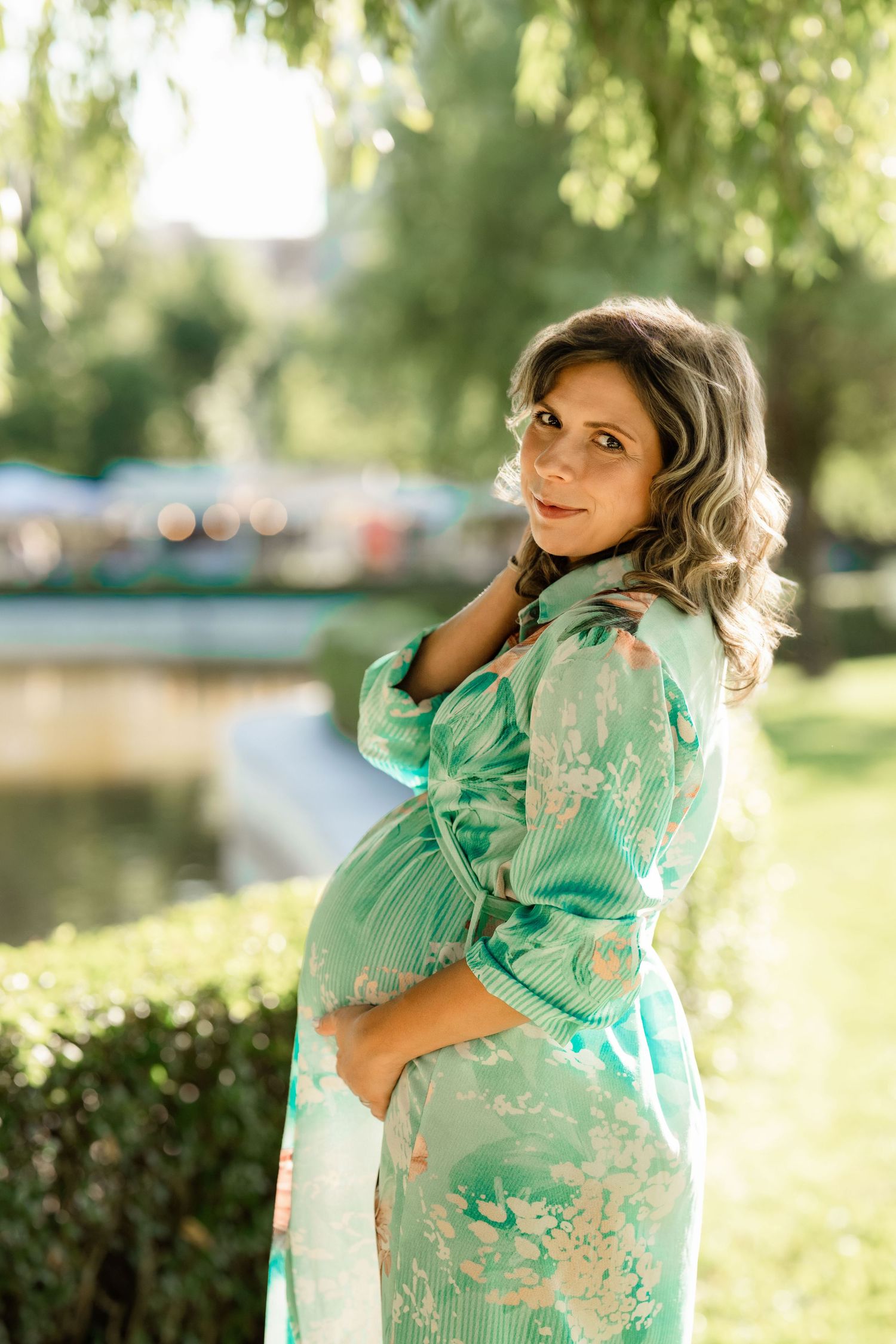 Outdoor maternity portrait of an expectant mother in a teal floral dress beside a pond on the Main Line, PA.