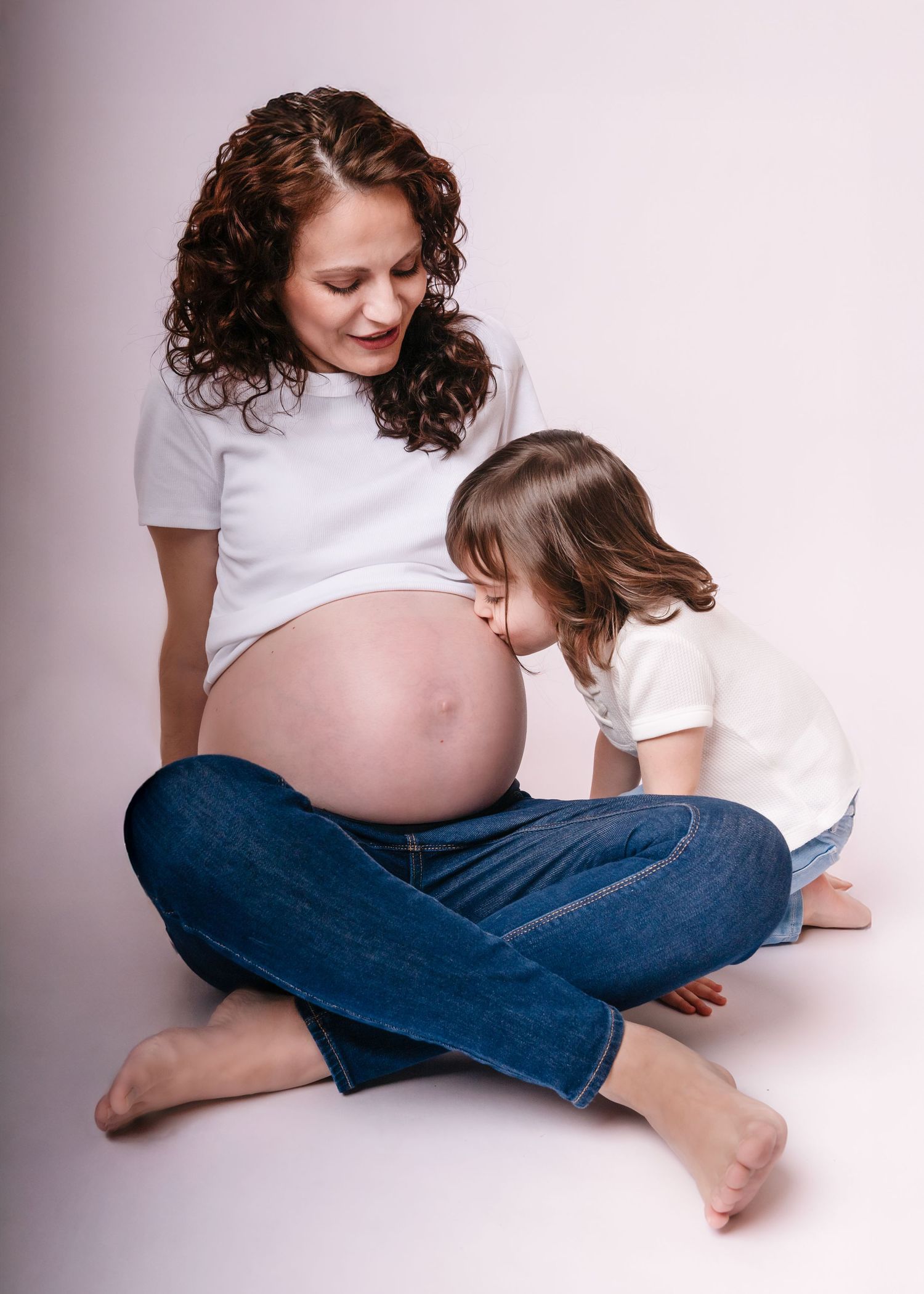 Pregnant woman in white tee and jeans sits cross-legged while young child kisses her bare baby bump.