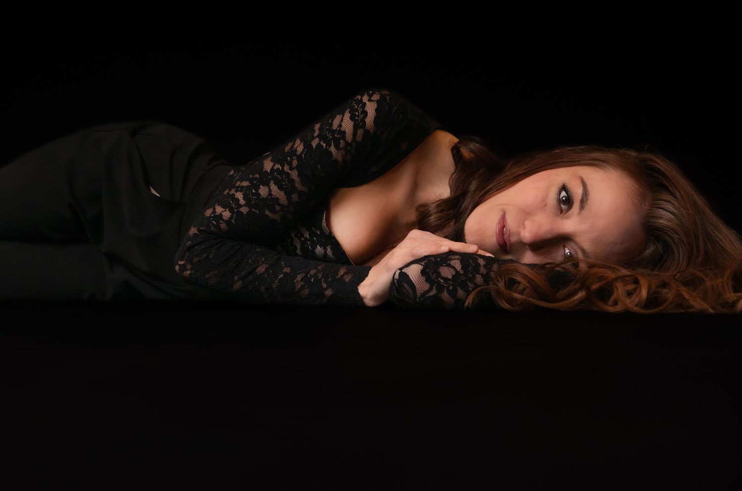 A woman in a black lace top lies gracefully on a dark surface, gazing at the camera with soft lighting.