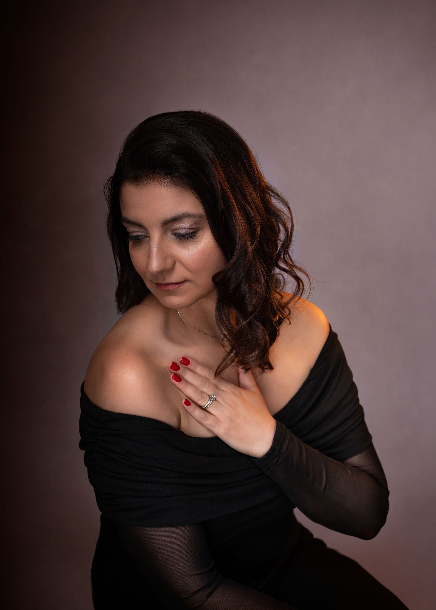 Woman in black off-shoulder top poses gracefully with red nails and ring against warm studio backdrop.