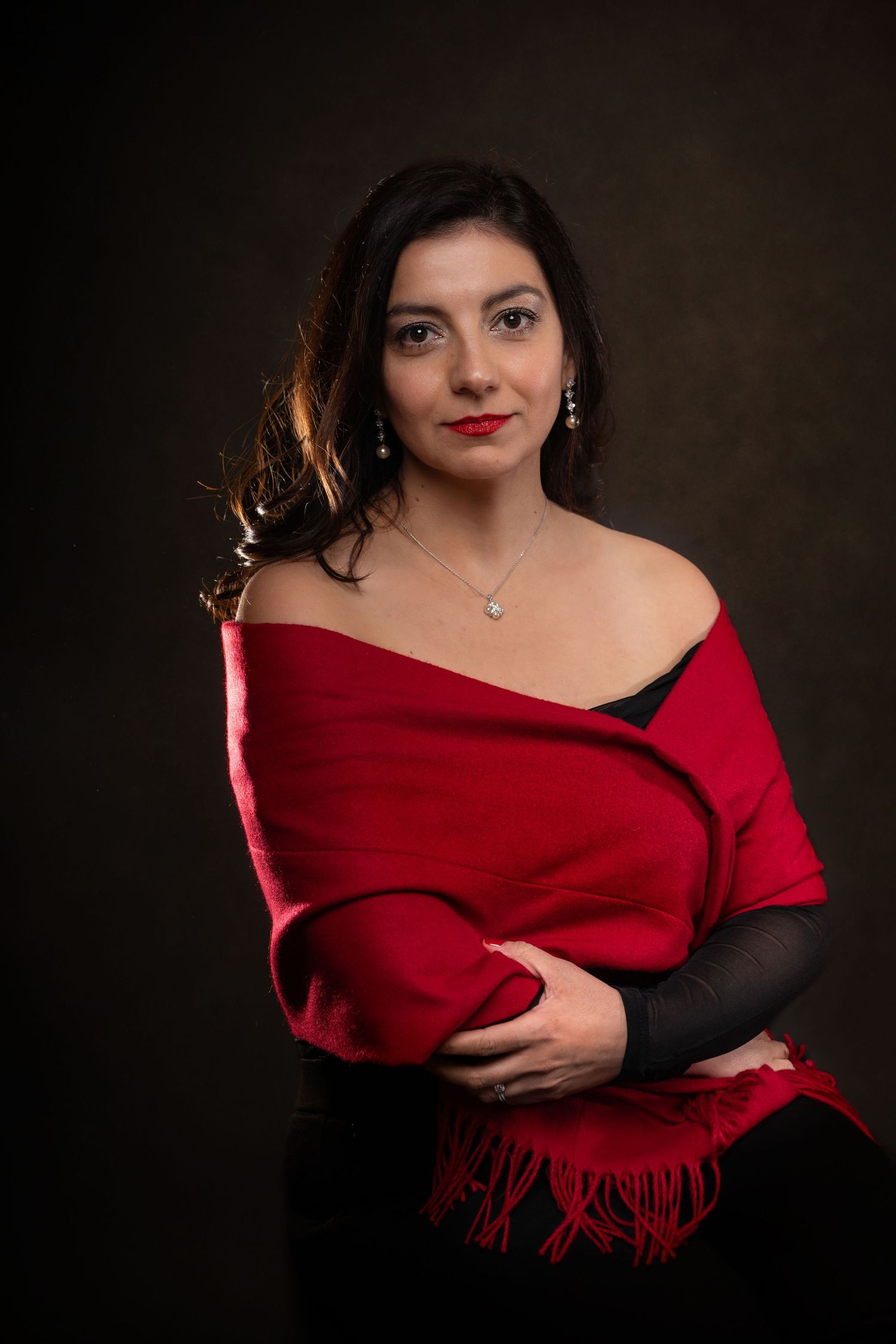 Woman in red shawl and black dress with jewelry poses elegantly against dark studio background.