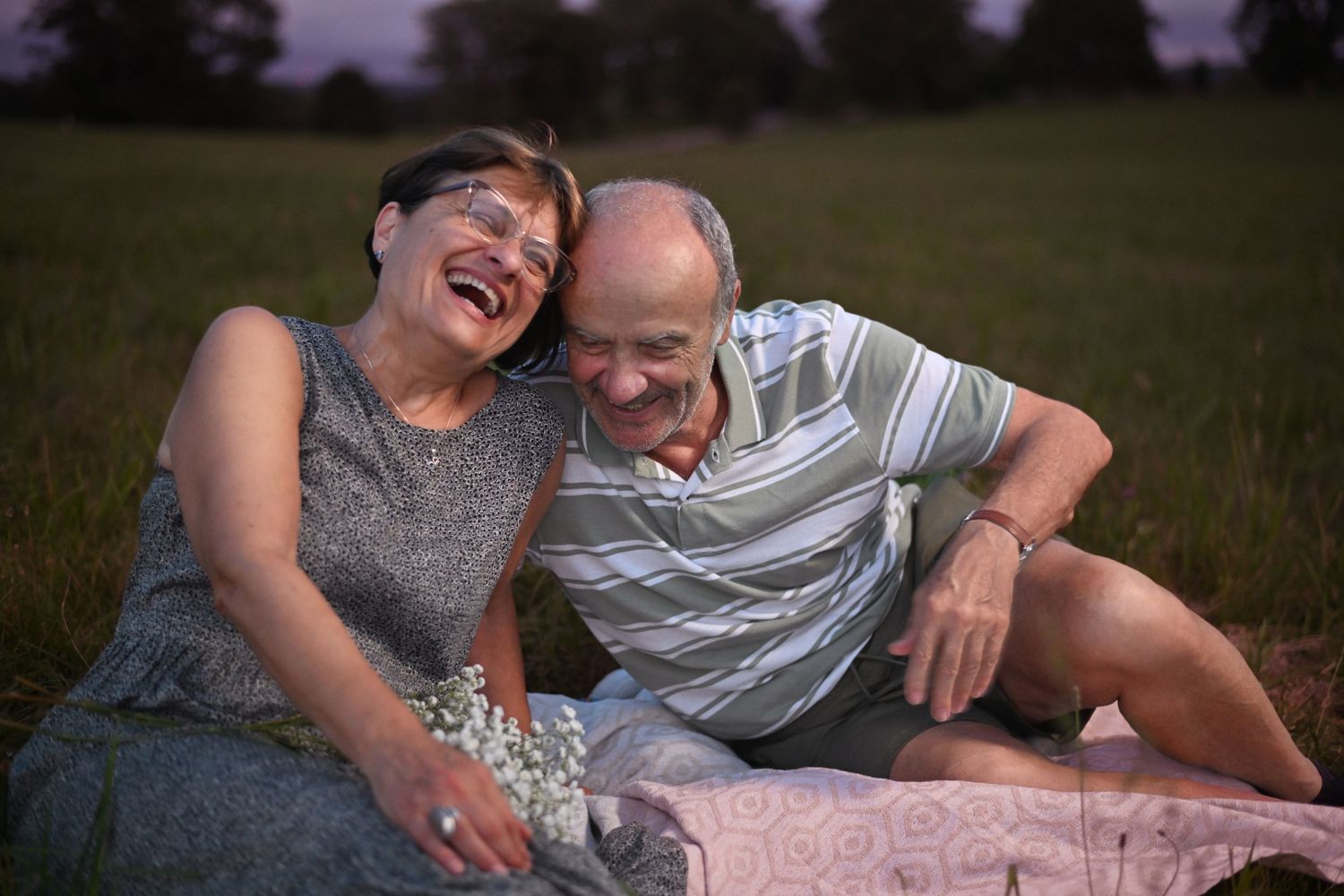 Playful couple laughing while lying on a blanket outdoors during a sunset photoshoot.