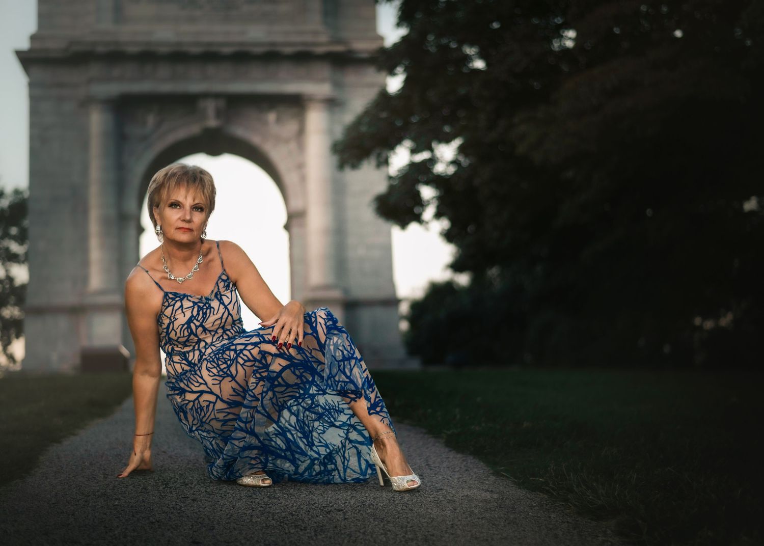 Woman in blue floral maxi dress poses on path before grand stone arch monument at dusk.