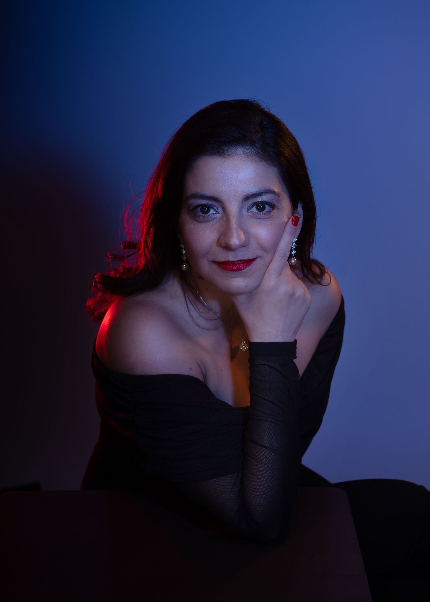 Woman in black off-shoulder top with red lips poses gracefully against a blue-lit background.