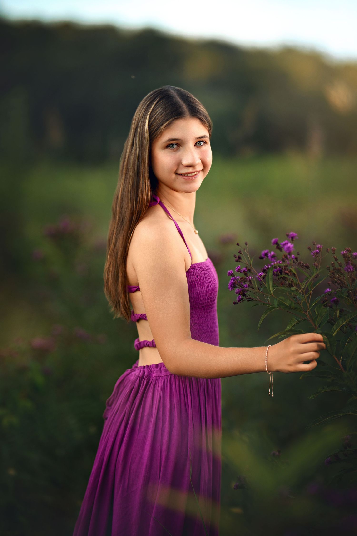 A young girl in a purple halter dress holds wildflowers in a sunlit green field.