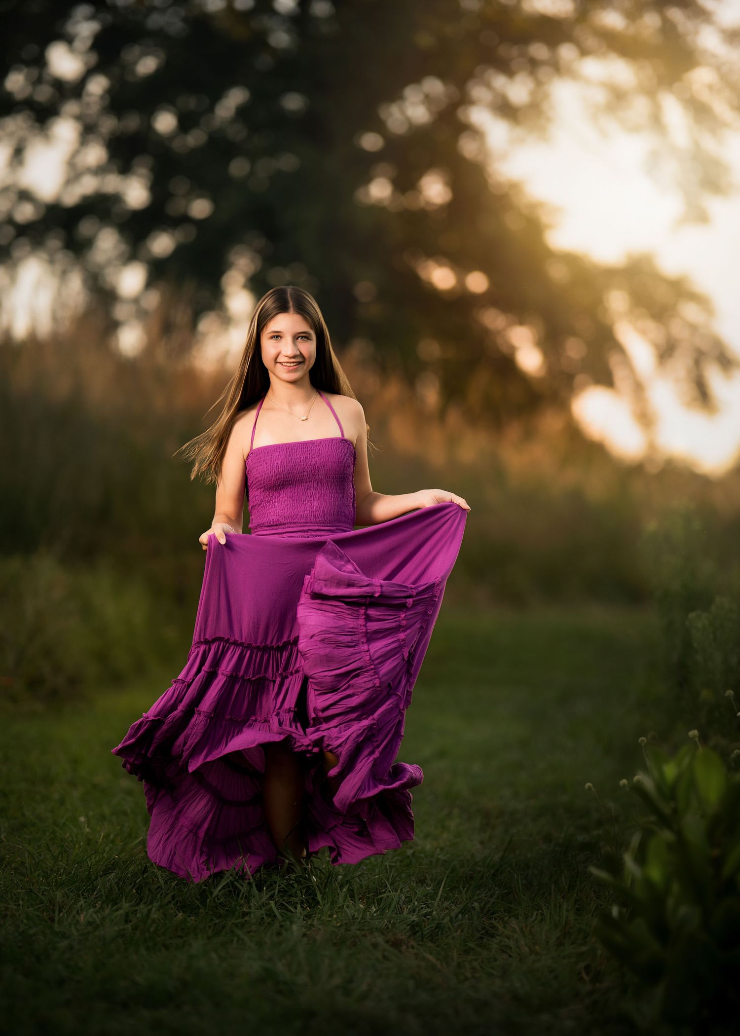 Young woman in a flowing purple halter gown poses outdoors at golden hour in a lush green field.