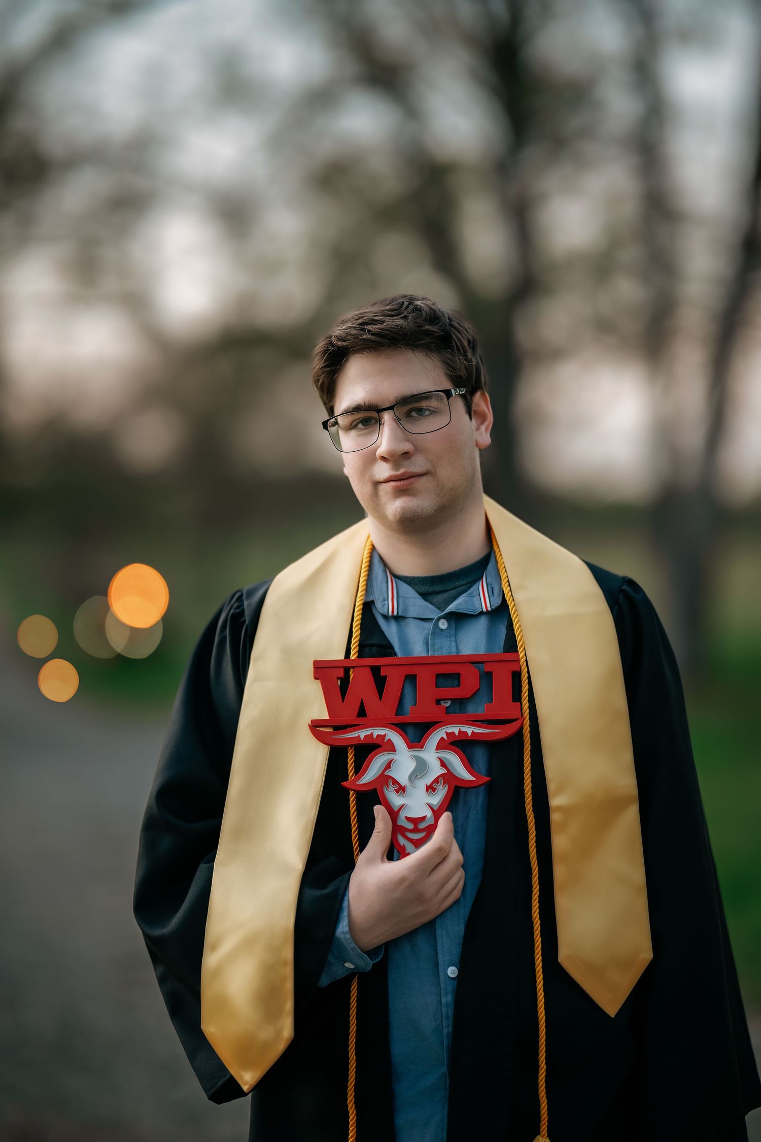 WPI graduate in black gown and gold stole holds red WPI logo sign outdoors at dusk.