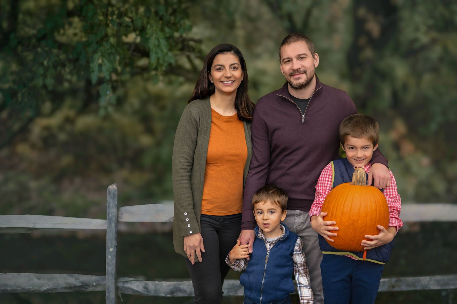 Family of four posing outdoors near a wooden fence in fall, with boy holding a large orange pumpkin.