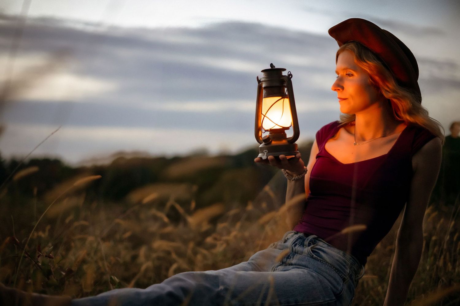 Woman in a red cap holds a glowing lantern while sitting in a golden field at dusk.