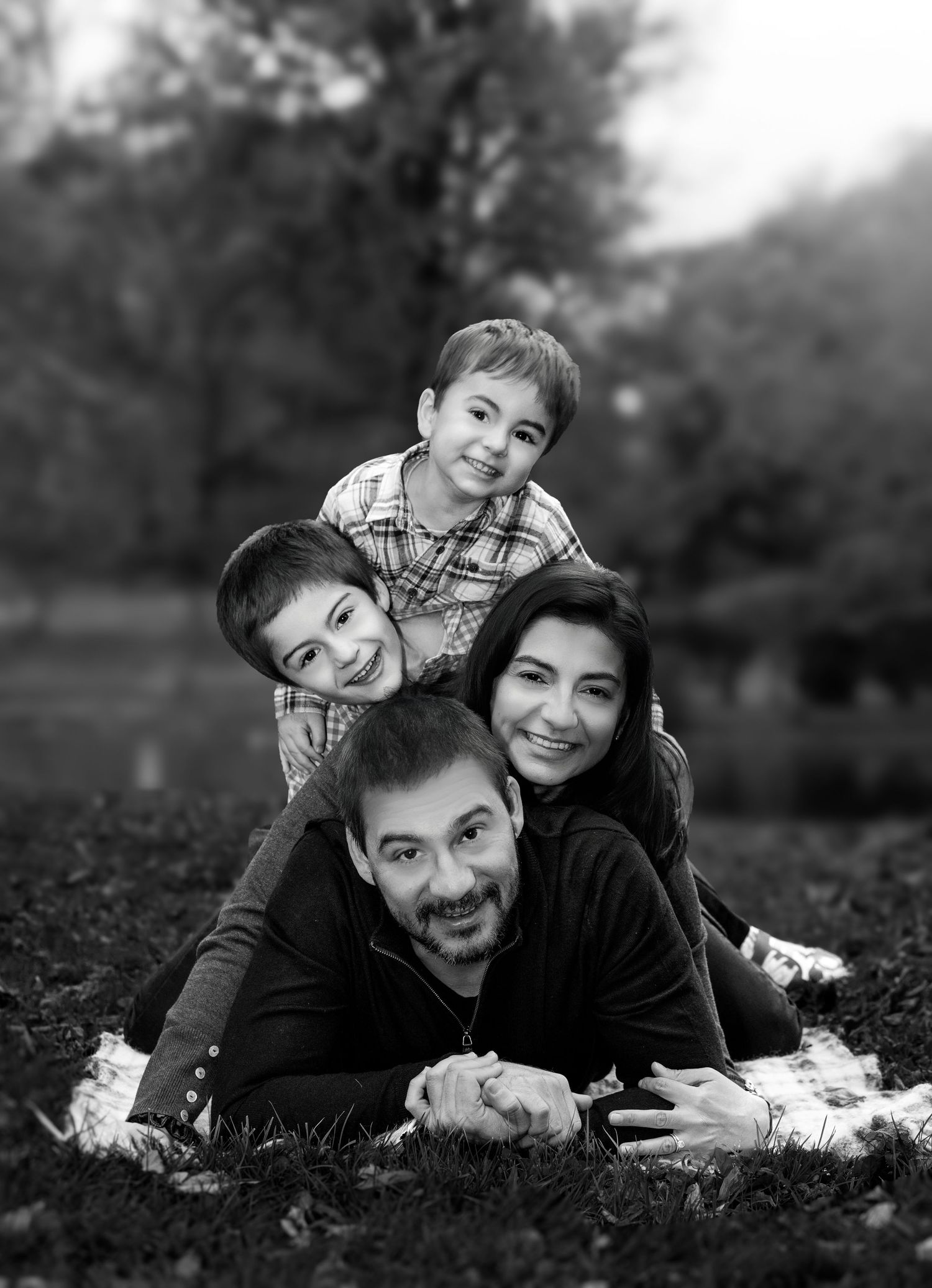 Black and white family portrait of parents and two young boys stacked together smiling outdoors on grass.