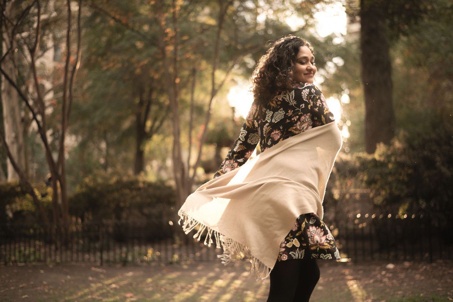 Woman in floral jacket twirling a cream fringed shawl in a sunlit autumn park.