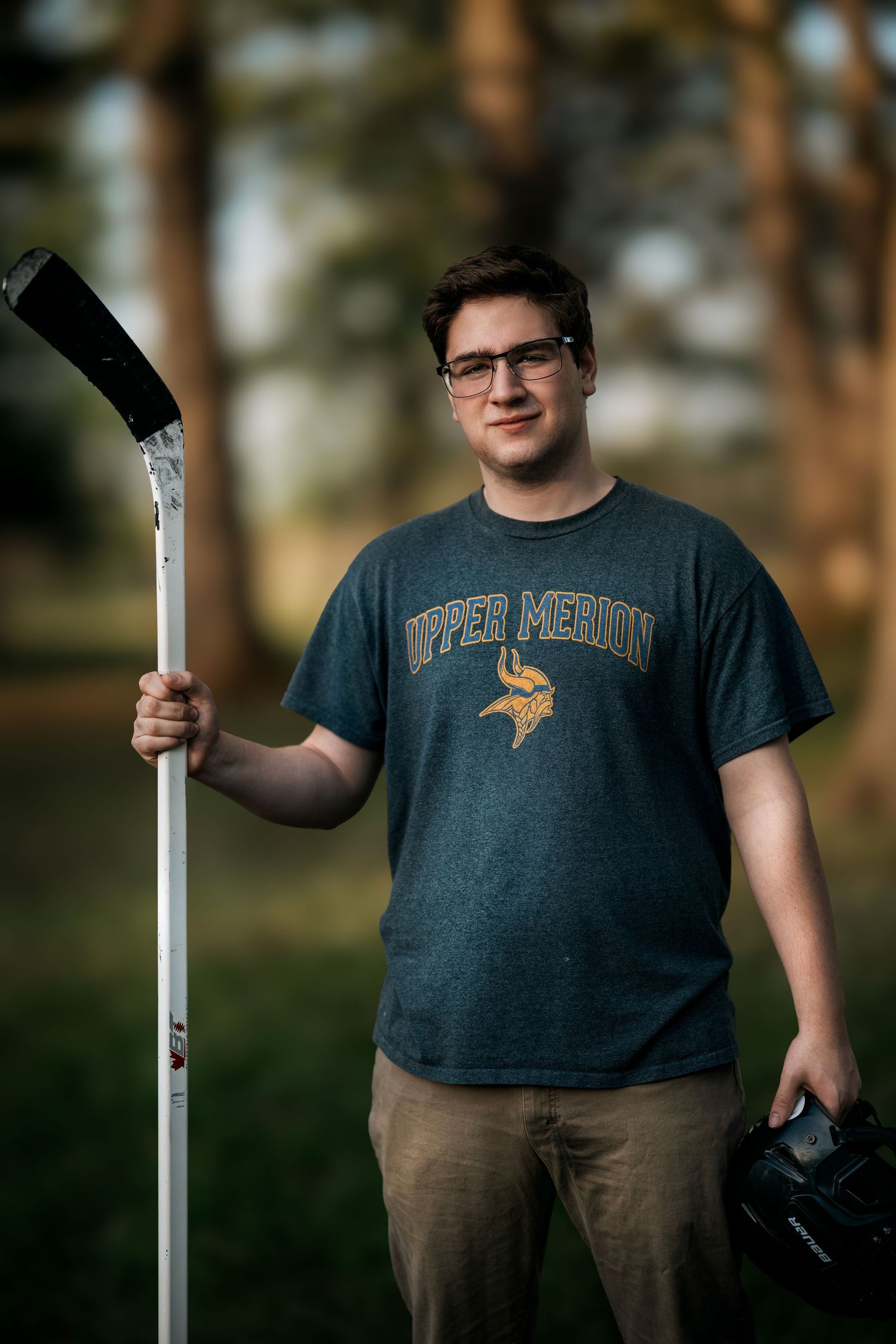 Young man in Upper Merion Vikings shirt holding hockey stick and helmet outdoors among trees.