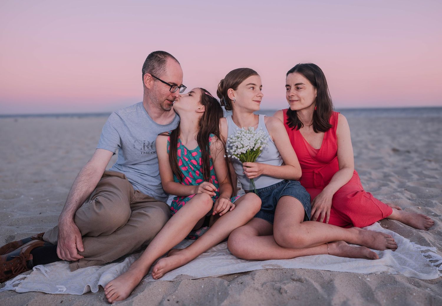 Family of four sitting on a blanket at the beach during sunset, with two girls holding flowers between parents.