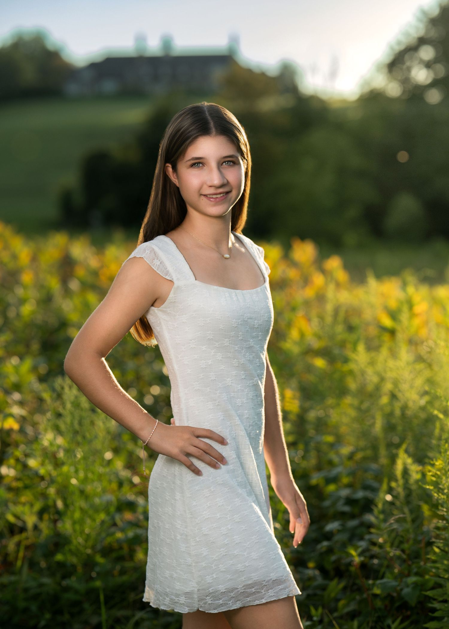 Young woman in white dress posing in golden field at sunset with green hills in background.