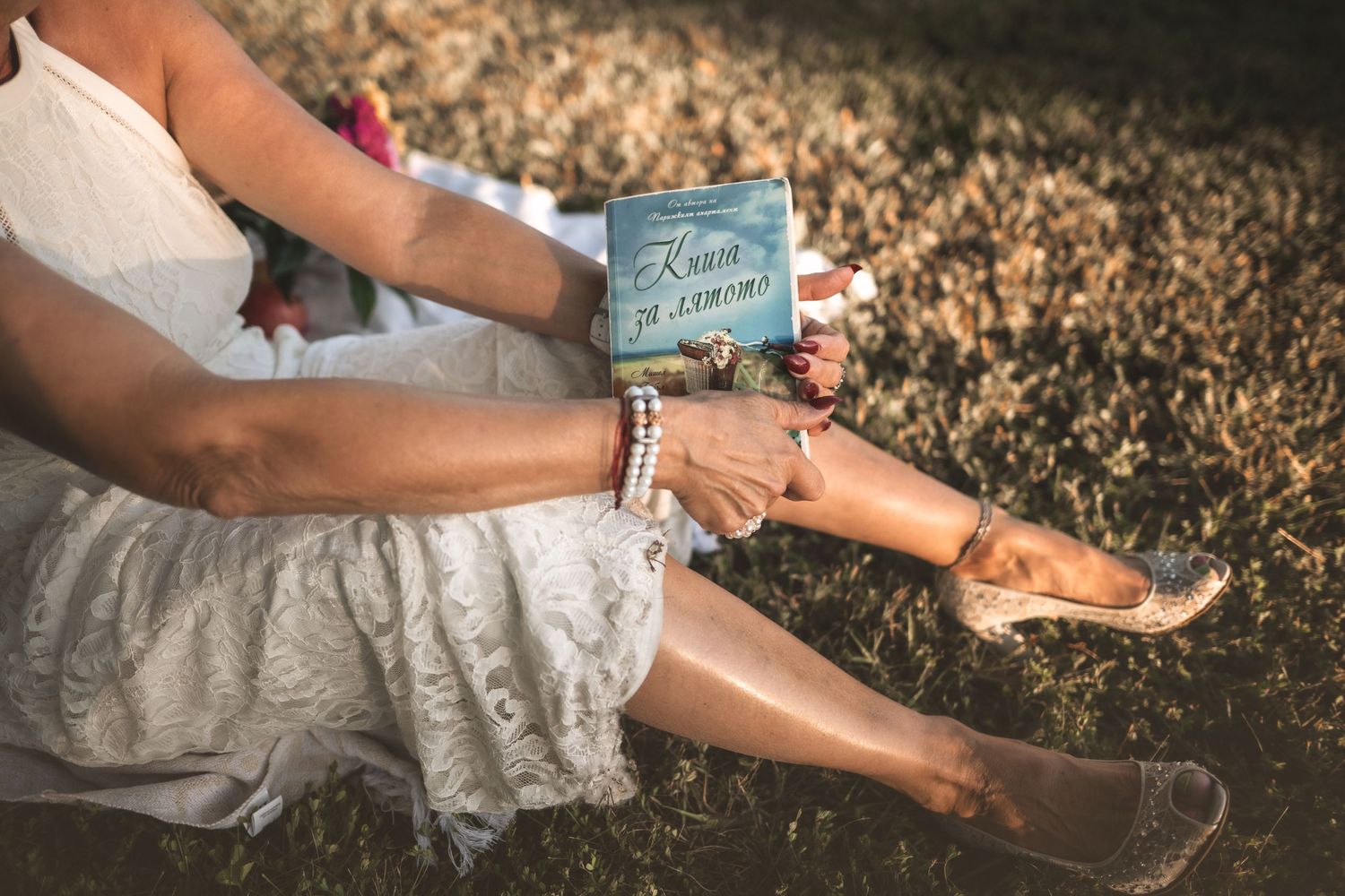 Hands holding a vintage-style book while sitting on a lace dress in warm sunlight.