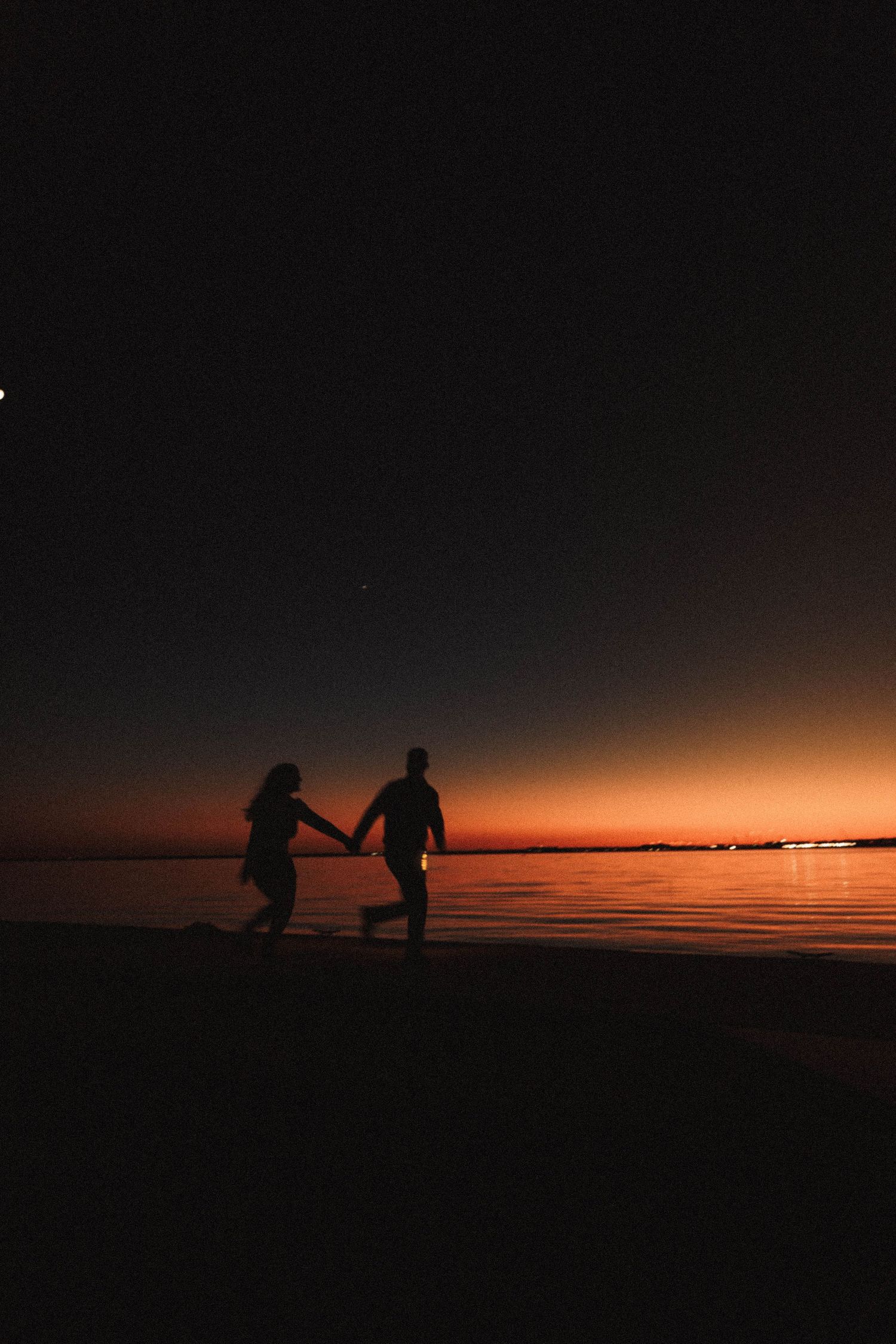 Two silhouettes hold hands and run along a beach at sunset with an orange sky reflecting on calm waters.