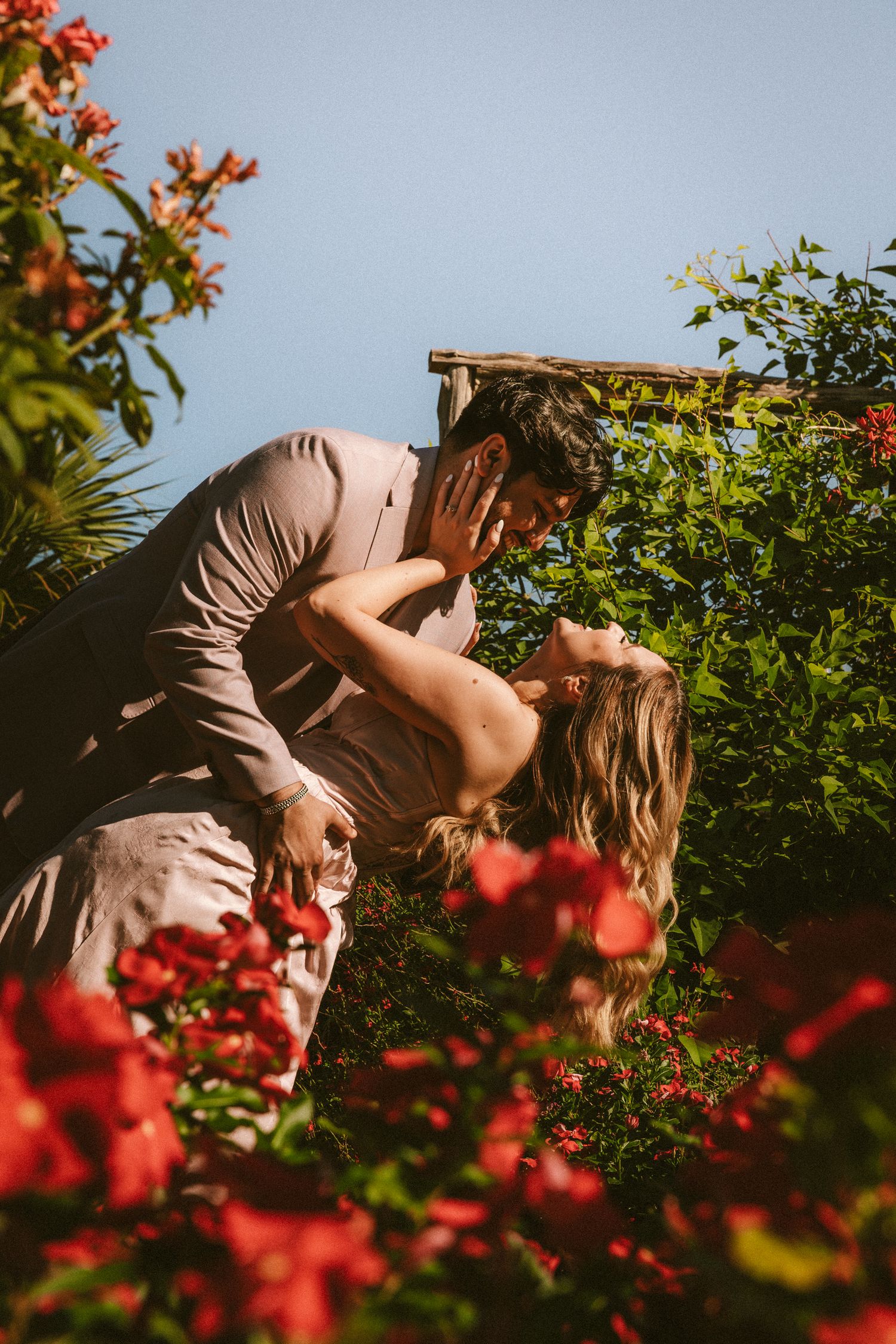 A romantic dip pose captured among vibrant red flowers in a garden setting.