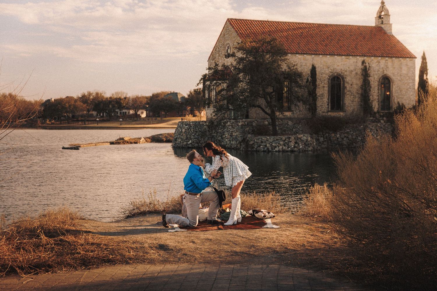 A romantic proposal scene at sunset by a stone church and lake, with a picnic blanket spread on the ground.