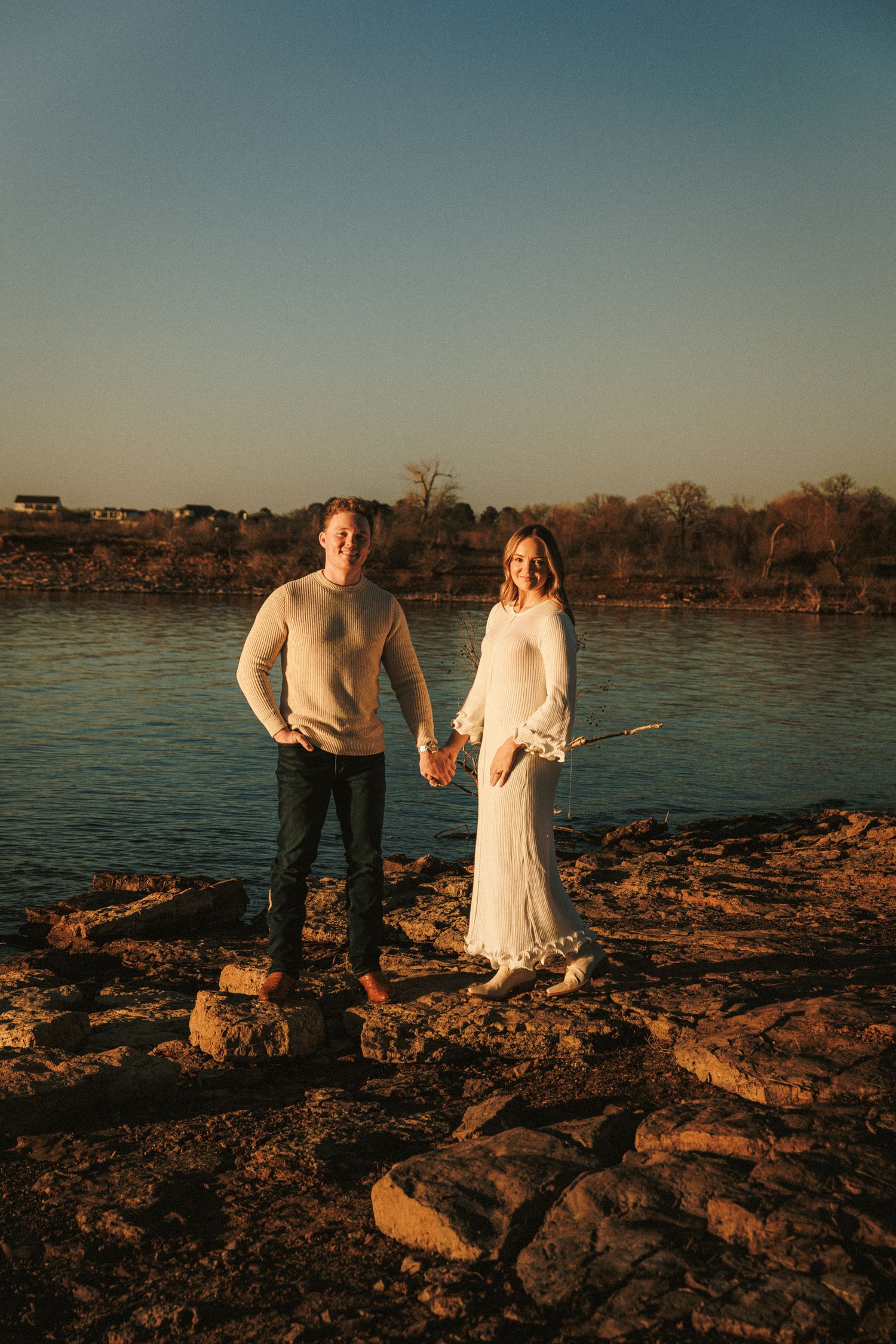 Couple in white and gray sweaters standing on rocky shoreline at sunset.