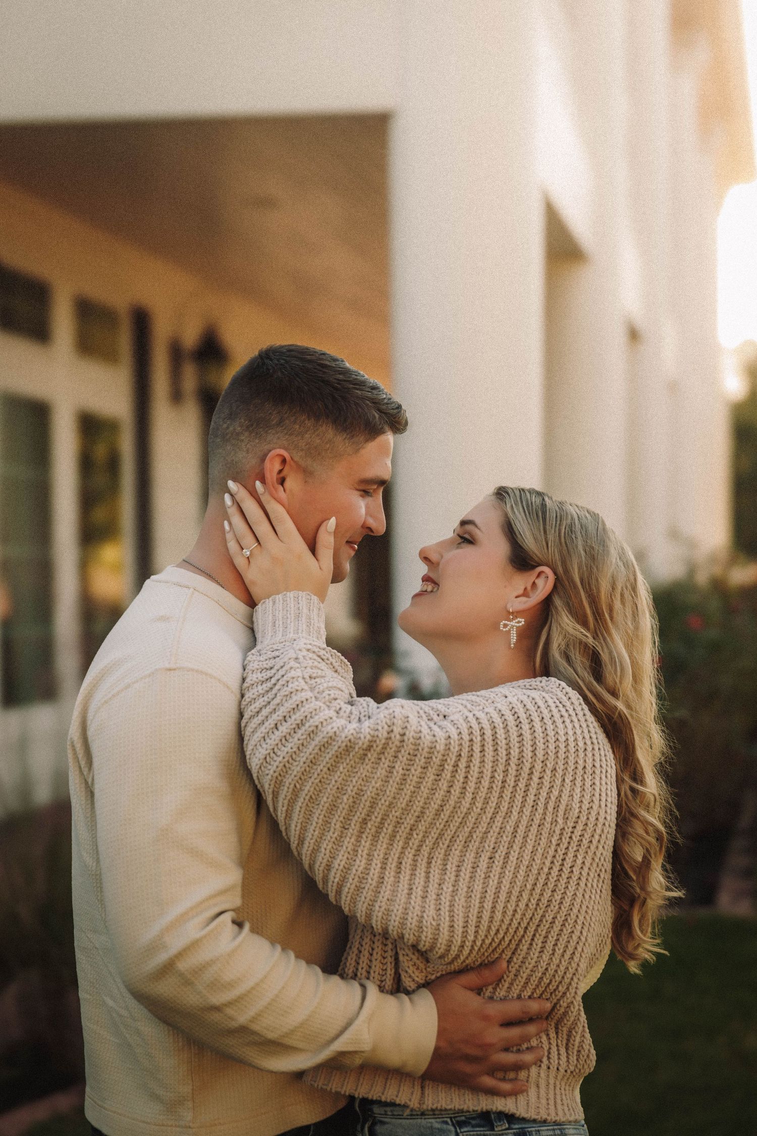 A romantic couple embraces outdoors at sunset wearing cozy cream sweaters against a neutral building wall.