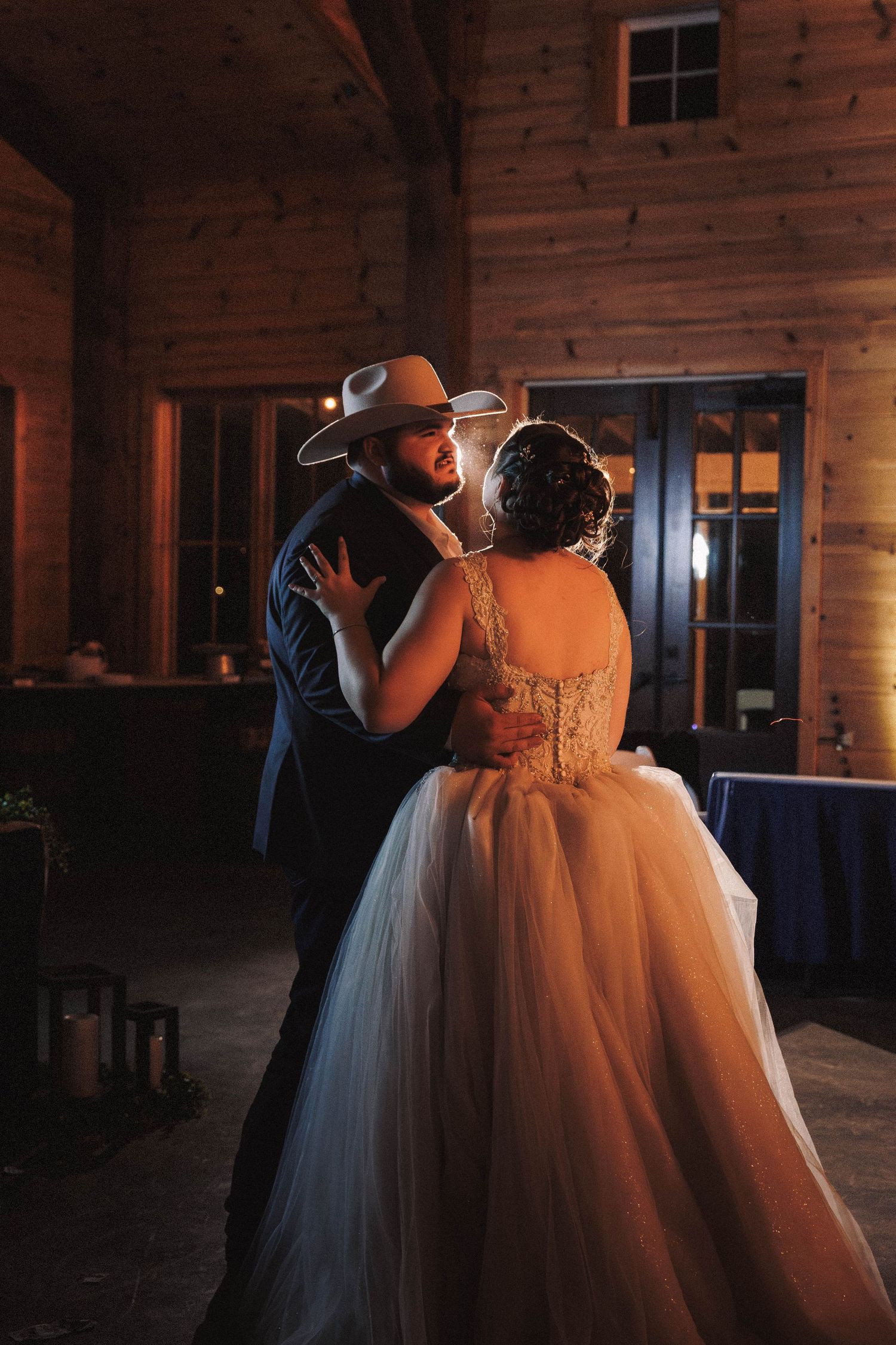 A couple shares their first dance in a rustic wooden barn venue illuminated by warm ambient lighting.