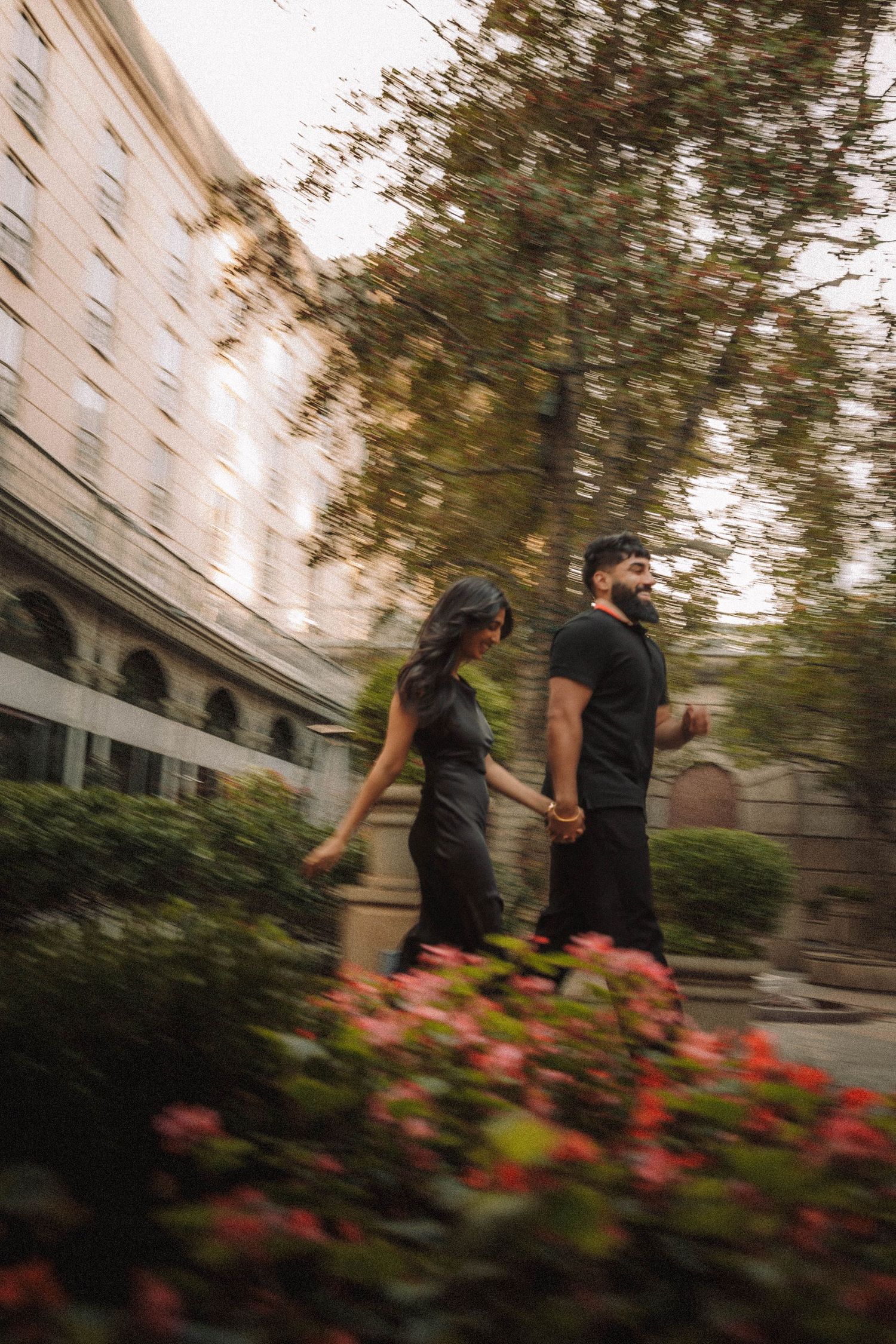 Blurred motion sequence of people walking past colorful flowers and buildings outdoors.