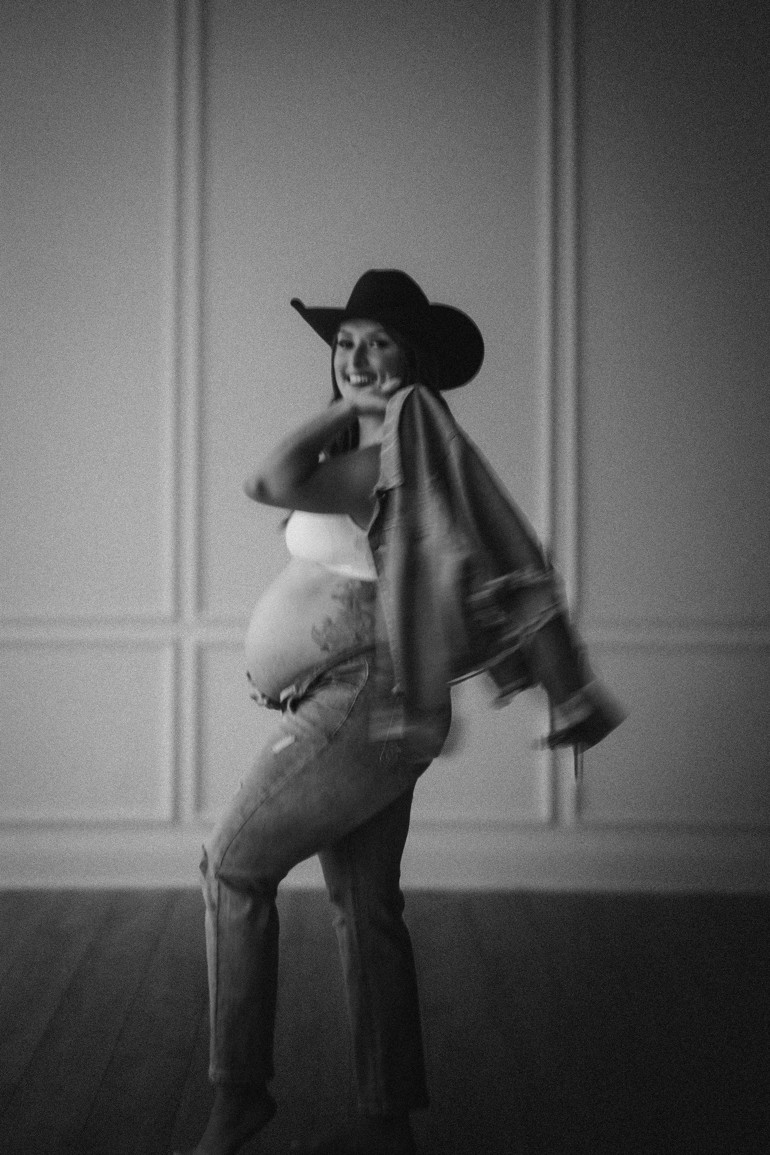 Black and white portrait of an expecting mother wearing a cowboy hat, cape and jeans in a minimalist studio setting.