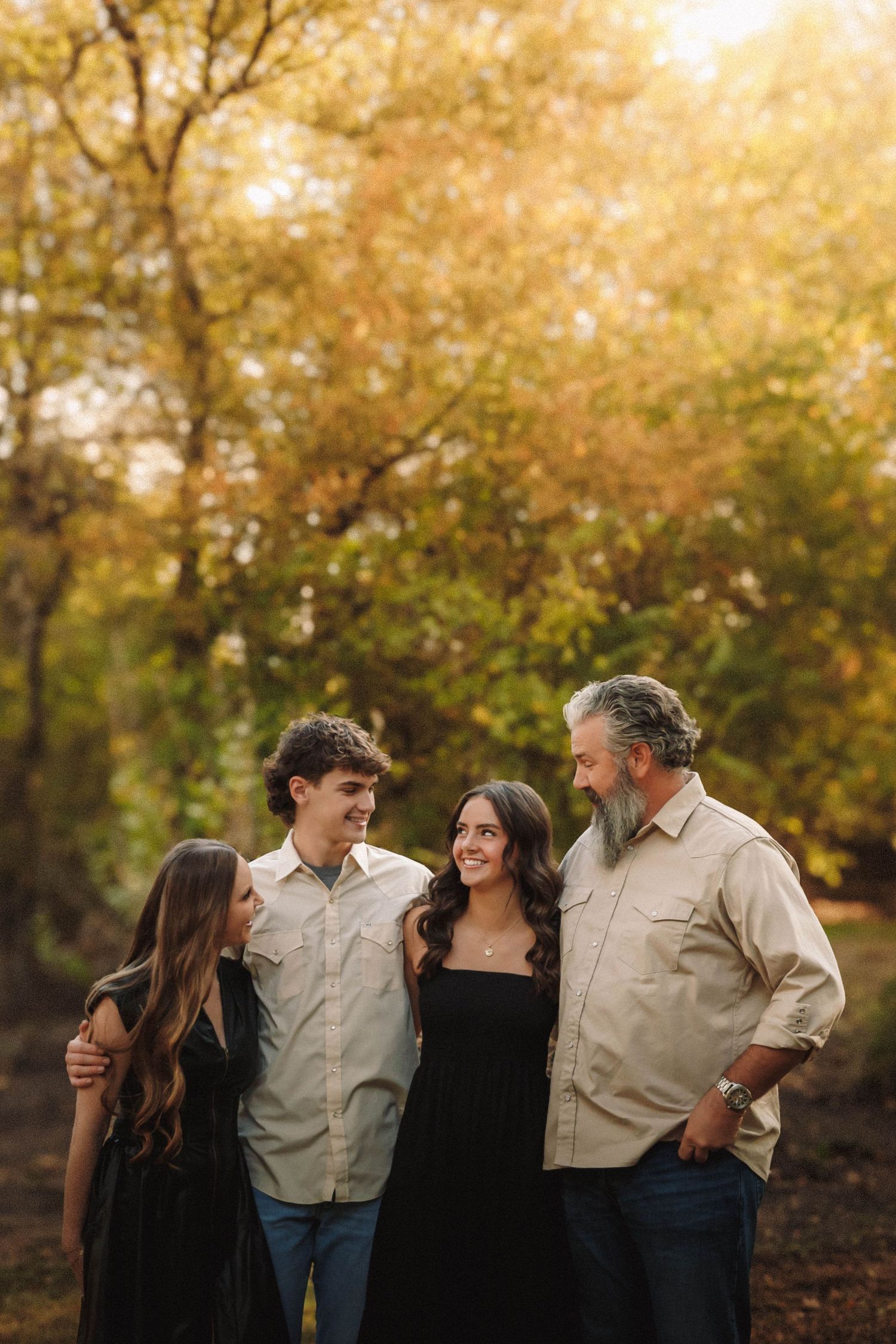 A group of people laugh together while posing for photos in a beautiful fall setting with golden trees in the background.