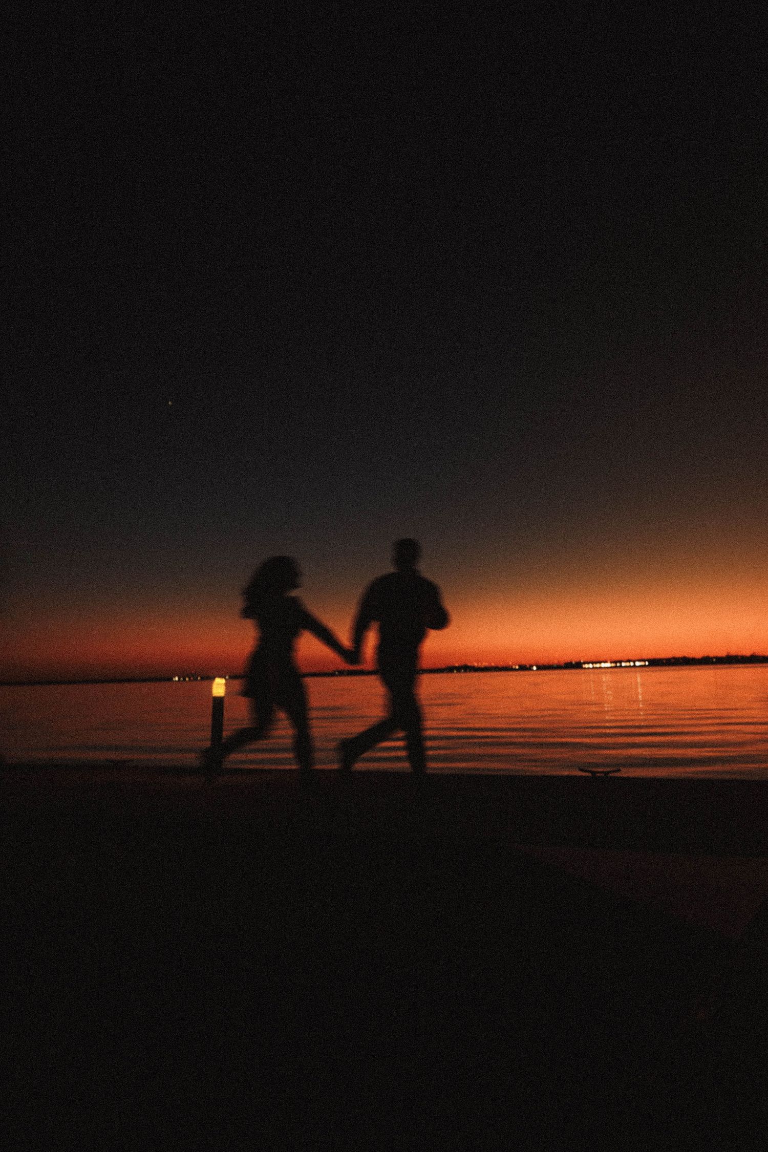 Silhouetted figures playfully interact against a dramatic orange sunset over water.