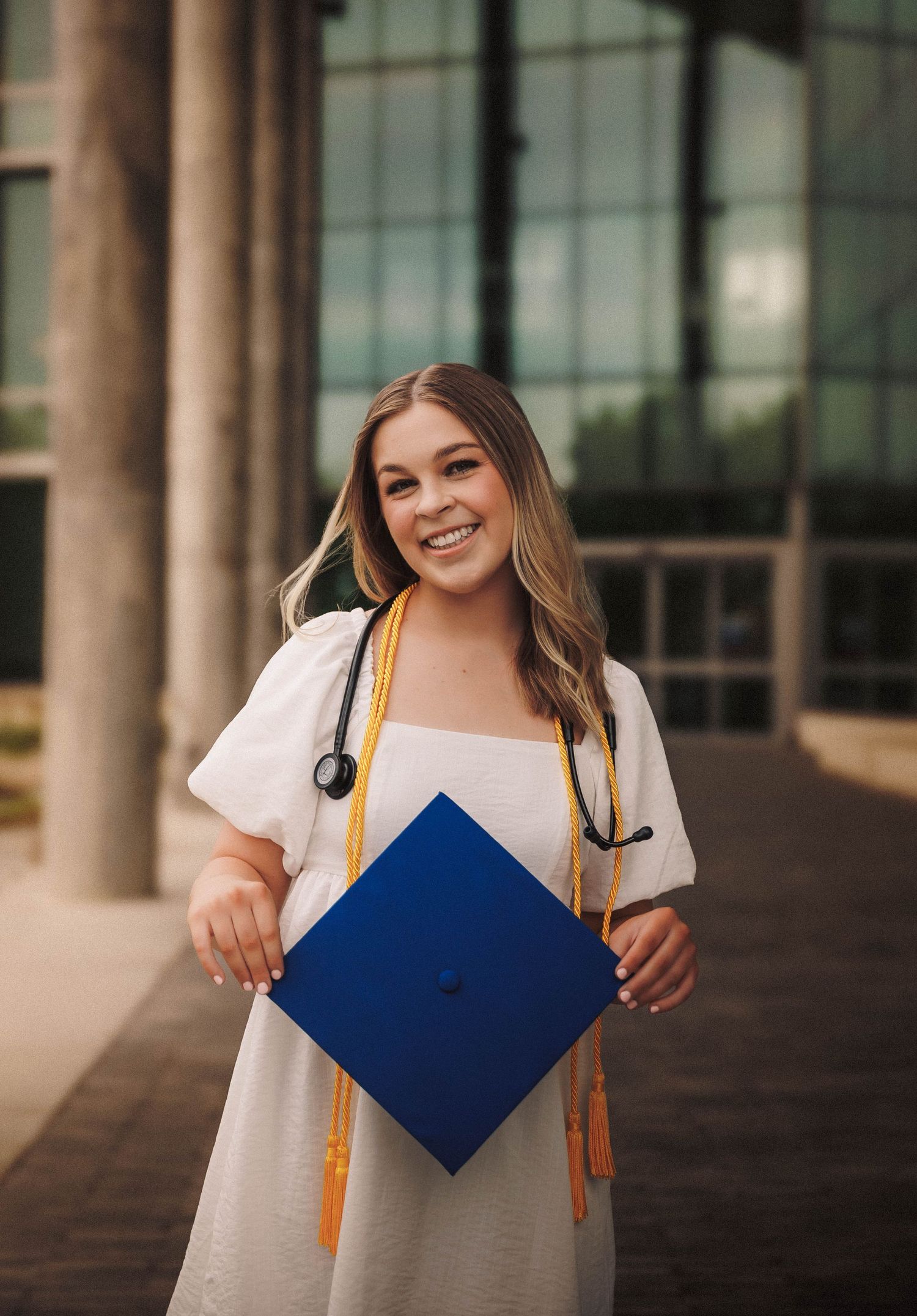 Graduate in white blouse holding blue diploma cover while posing between architectural pillars.