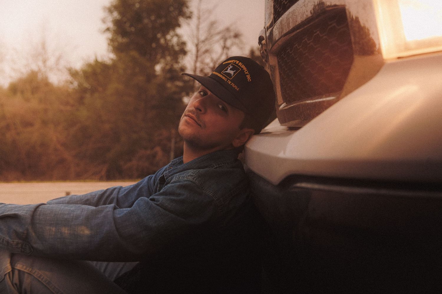 Someone wearing a baseball cap and denim jacket leans against a car during golden hour sunset with eyes closed.