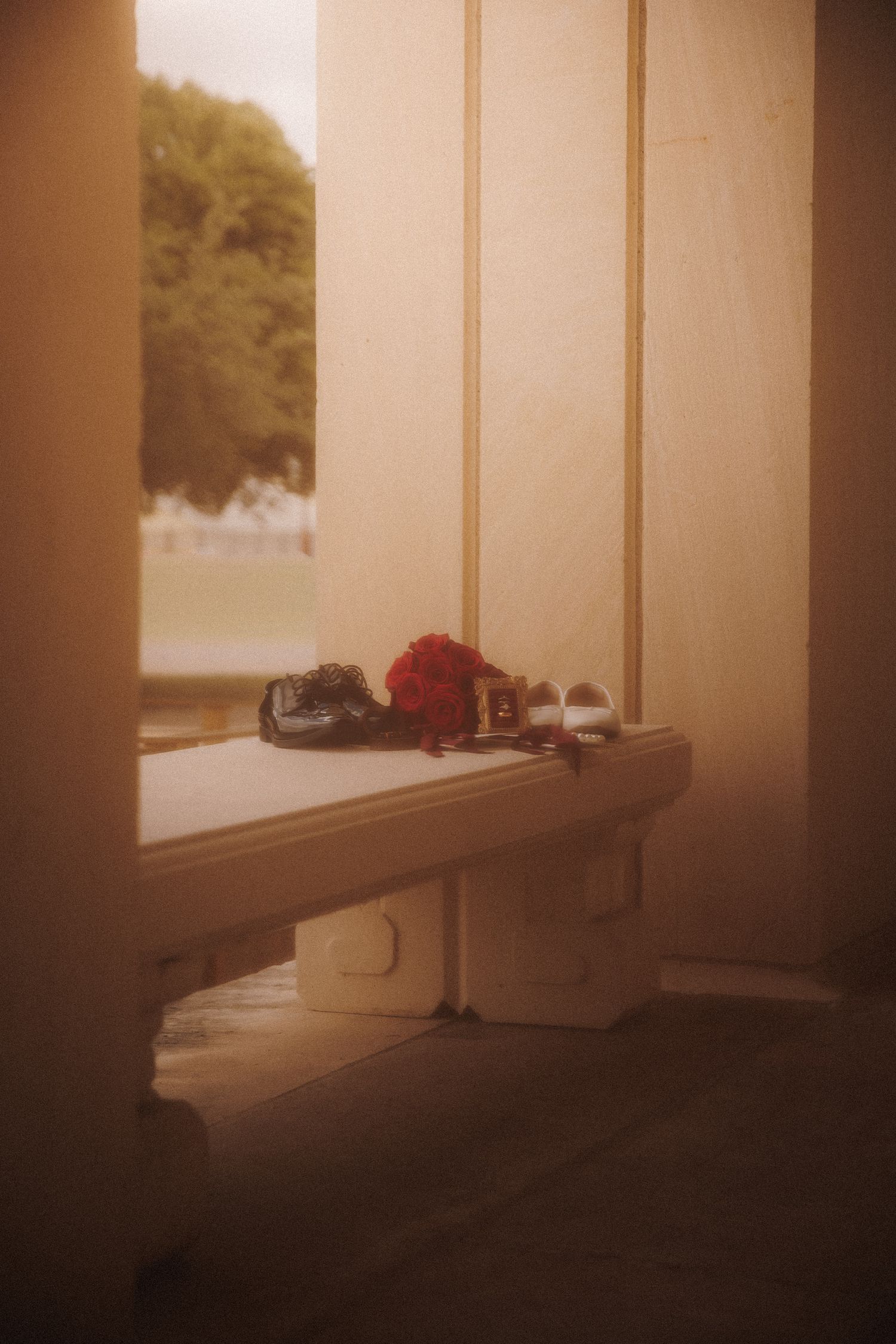 Vintage porch bench with red flowers and small shoes beside a window overlooking a green garden.