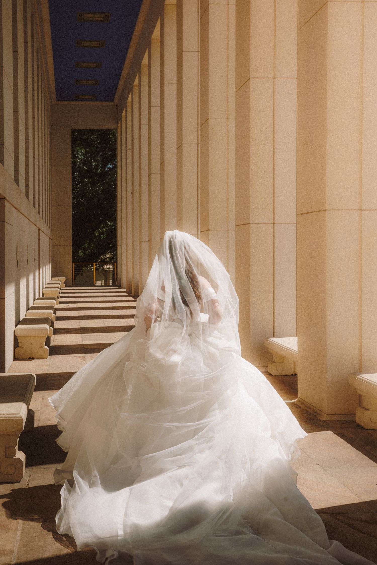 Bride in flowing white ball gown and veil walking between grand stone columns.