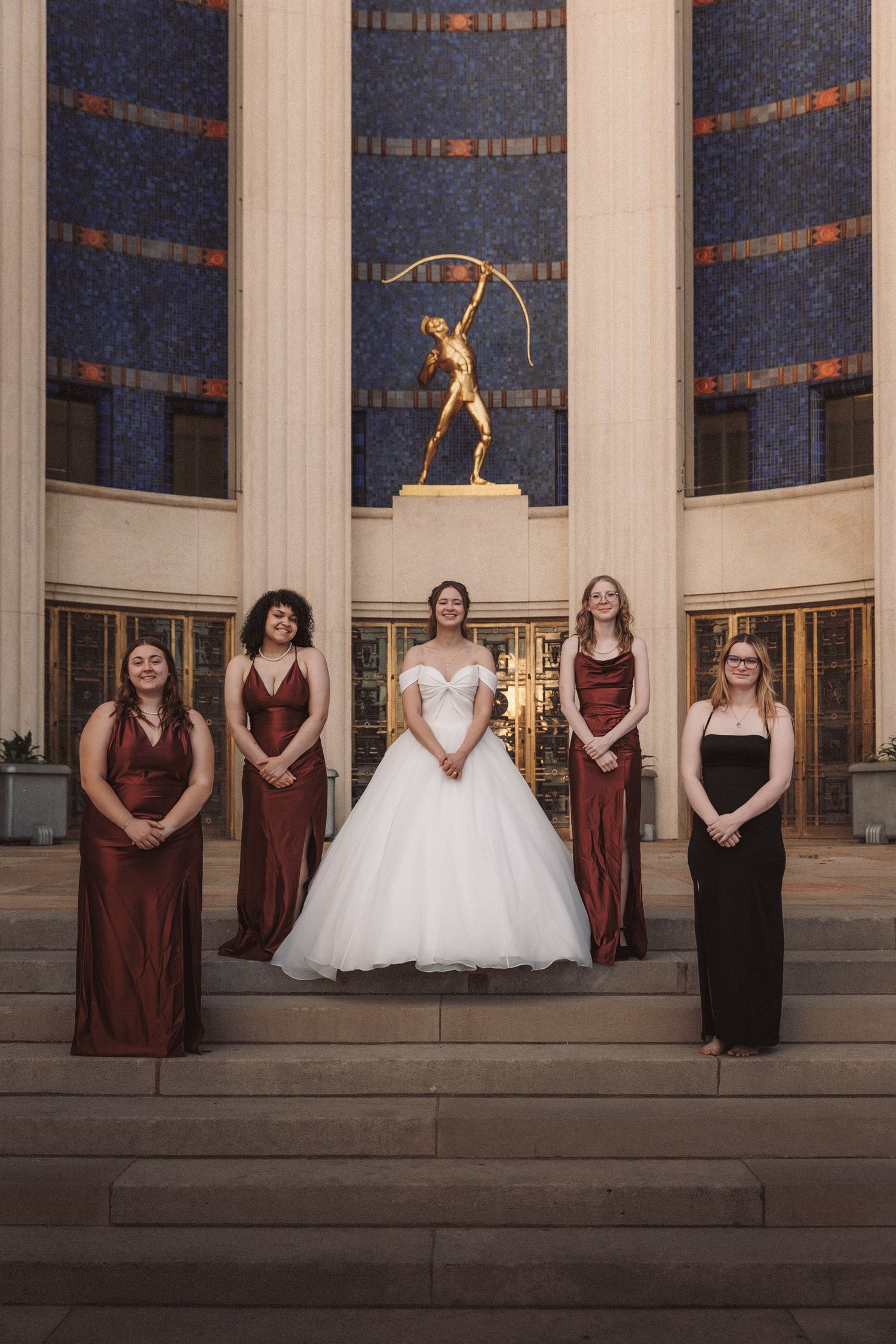 Bride in white ball gown with four bridesmaids in burgundy and black dresses on steps of a neoclassical building.