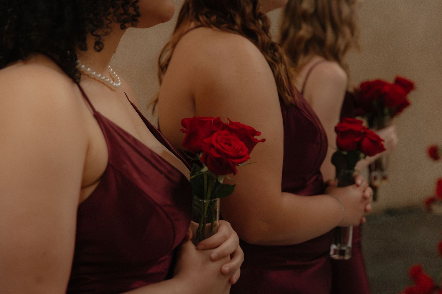 Bridesmaids in burgundy satin dresses holding single red roses at a wedding ceremony.