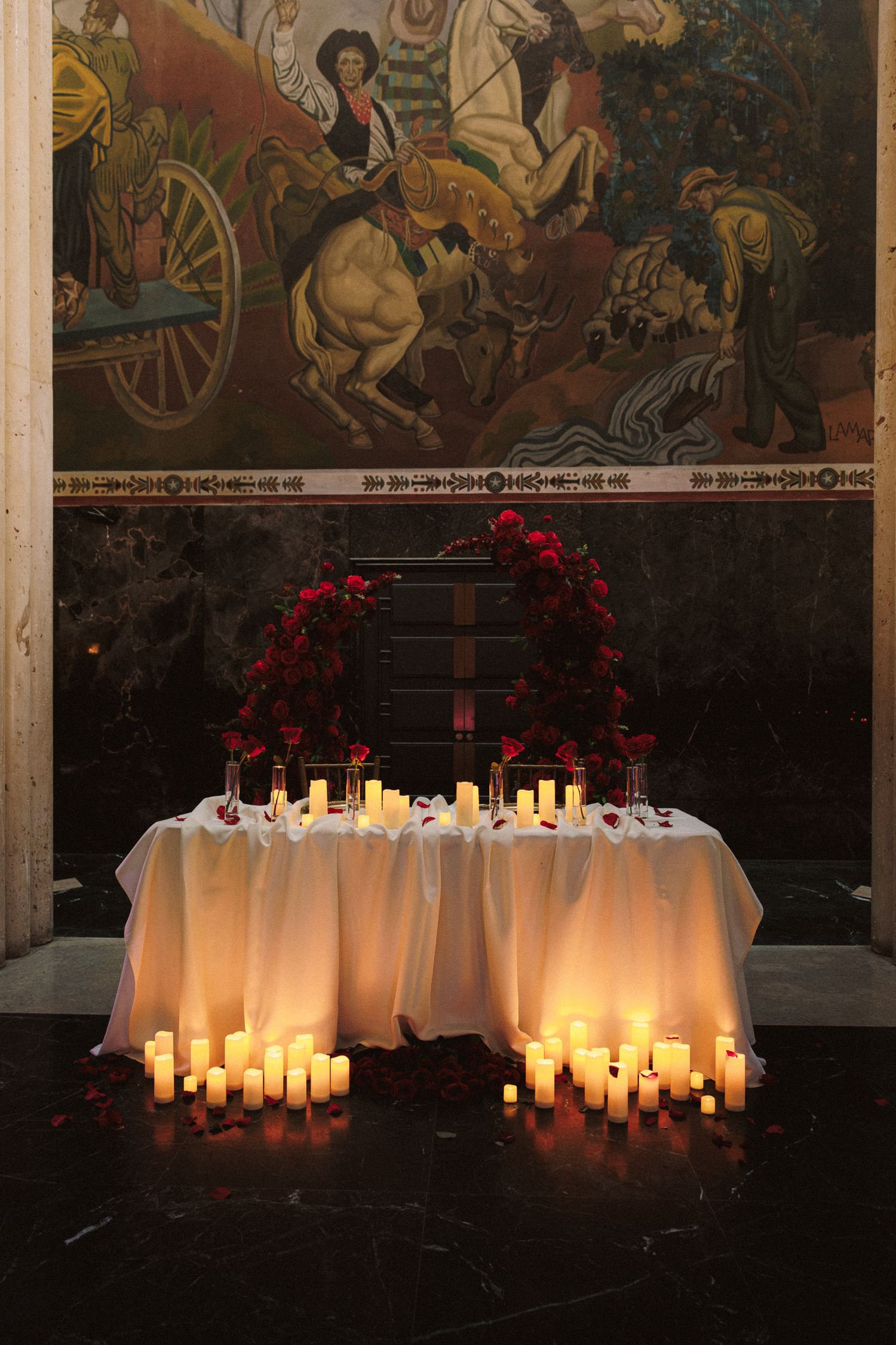 Romantic sweetheart table adorned with red roses, glowing candles, and white draping beneath a classic mural.