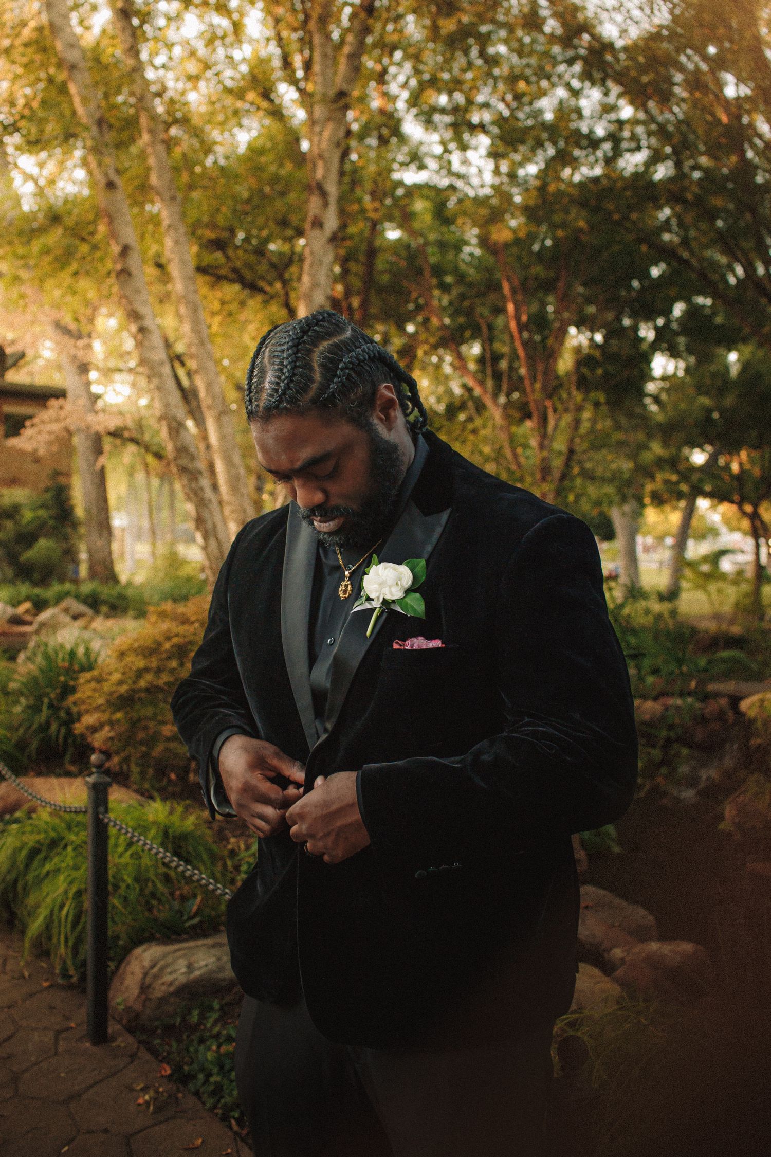Groom in black velvet tuxedo with white boutonniere adjusting jacket in autumn garden setting.