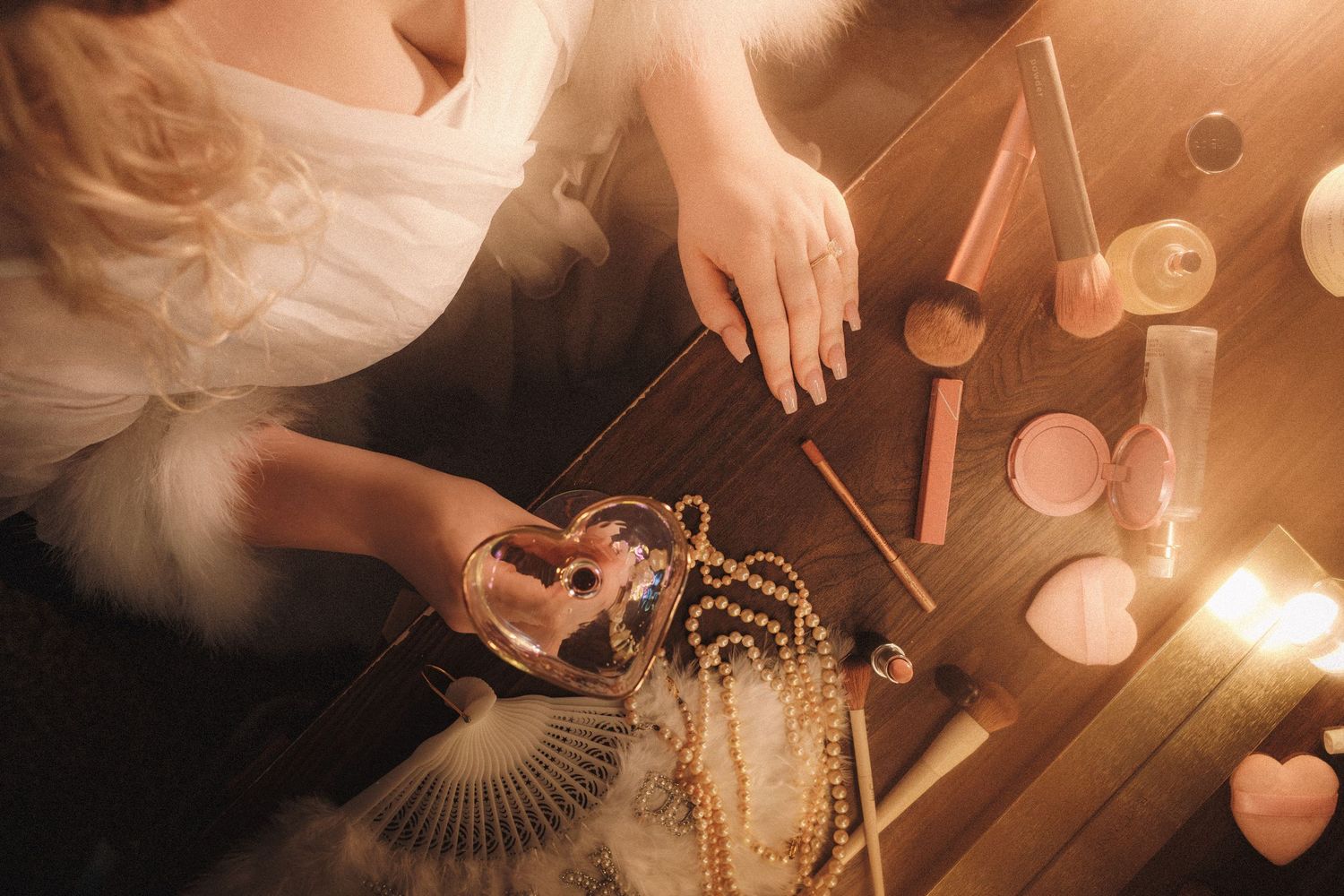 Overhead view of a bride at wooden vanity with pearl jewelry, makeup, perfume, and candles in warm candlelight.