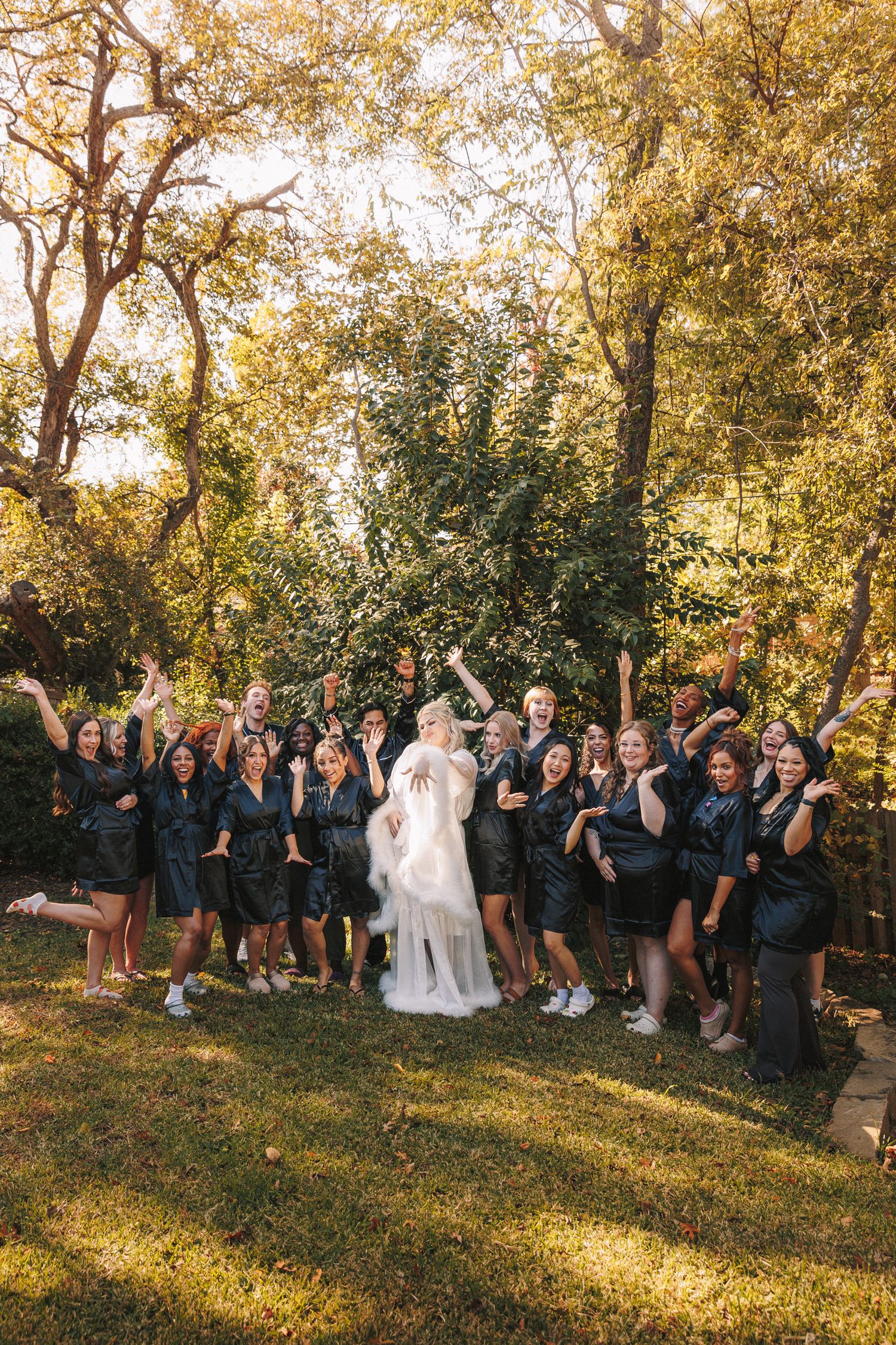 Bride in white gown celebrates with large bridal party in matching black robes outdoors among golden autumn trees.