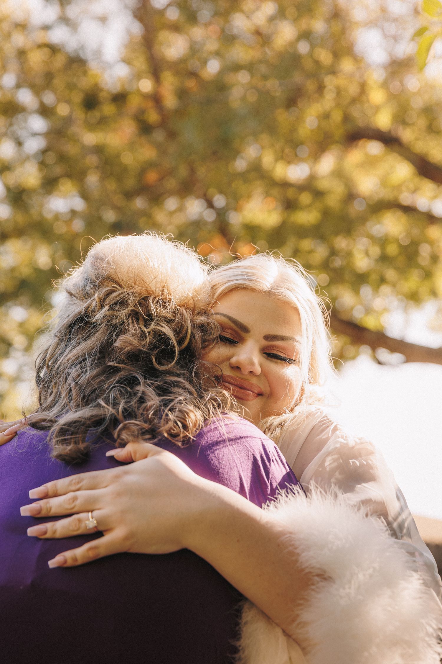 Smiling blonde bride in fur stole hugging guest outdoors under golden autumn trees.