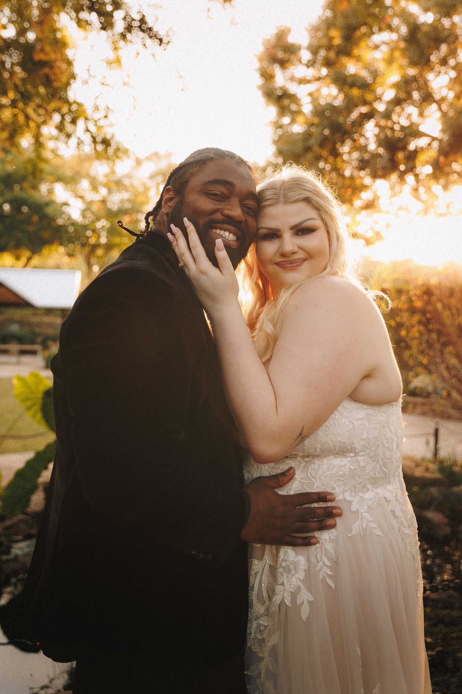 Interracial newlywed couple smiling and embracing in warm golden sunset light outdoors after wedding ceremony.